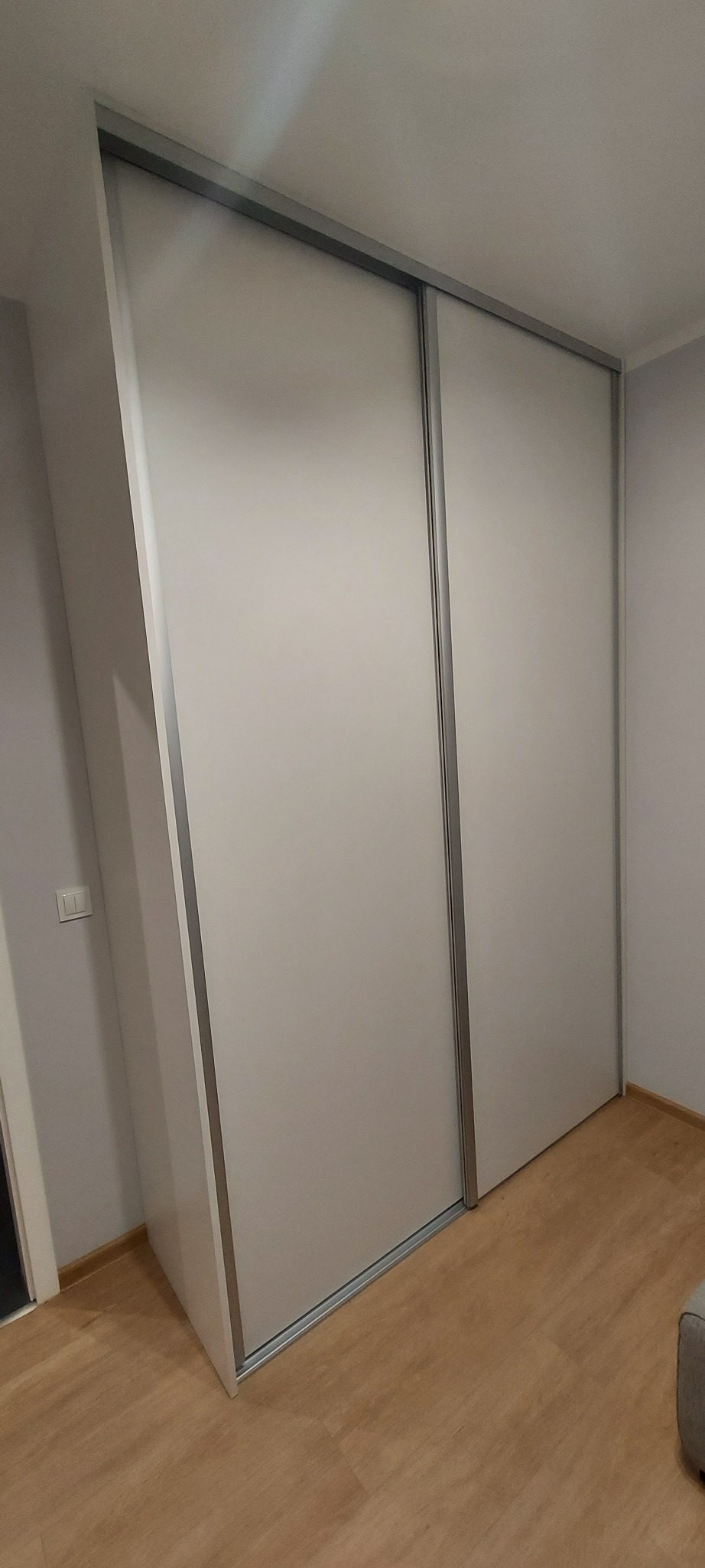 Biała szafa przesuwna z aluminiowymi profilami, dopasowana do skosu ściany, widoczna na tle jasnego pokoju z panelami podłogowymi.