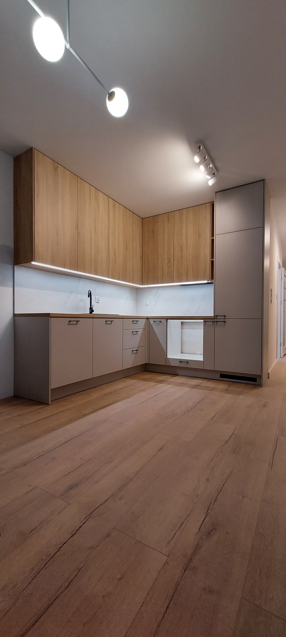Nowoczesna kuchnia z drewnianymi frontami szafek górnych i szarymi dolnymi, oświetlona listwą LED pod szafkami i lampą sufitową, z drewnianą podłogą.