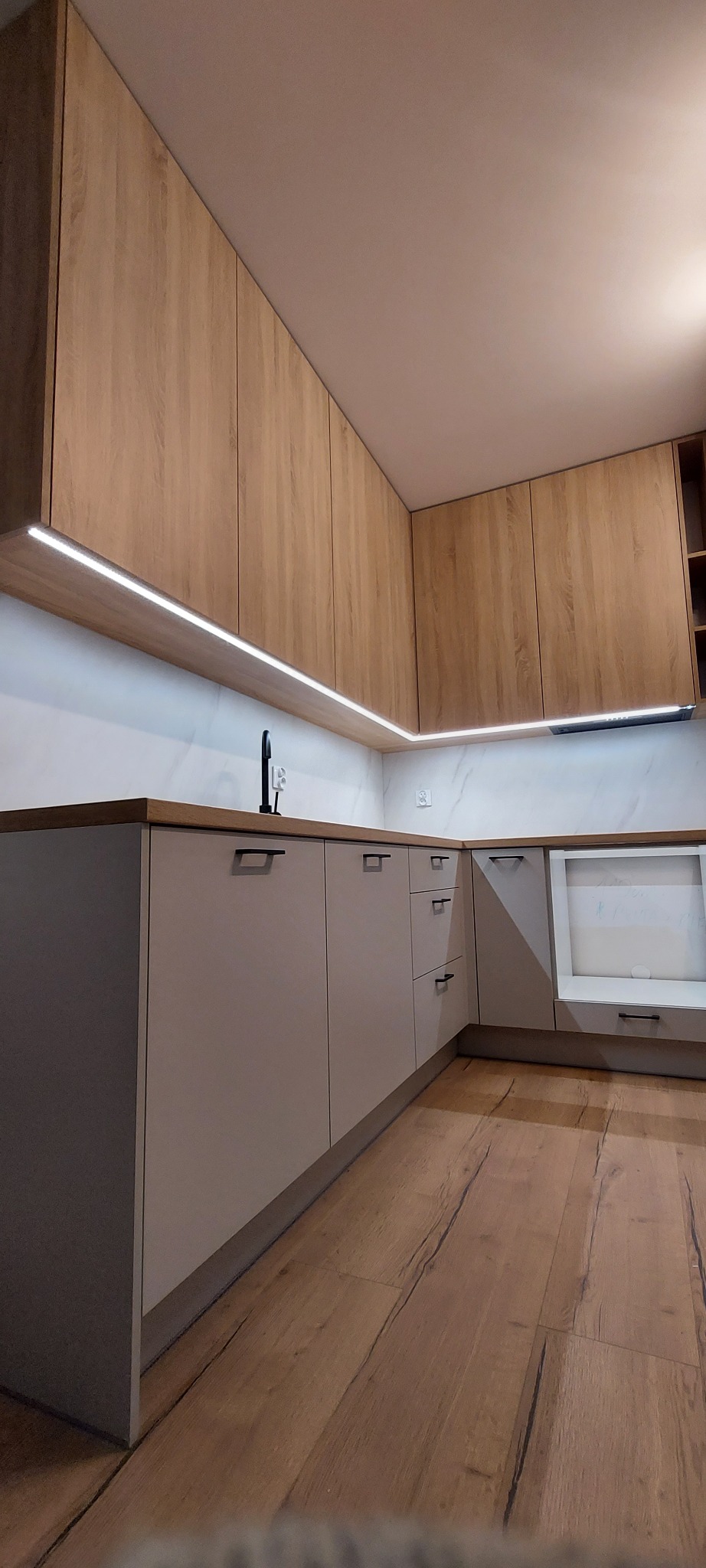 Nowoczesna kuchnia z drewnianymi górnymi szafkami z oświetleniem LED pod spodem i szarymi dolnymi szafkami z czarnymi uchwytami oraz drewnianą podłogą.