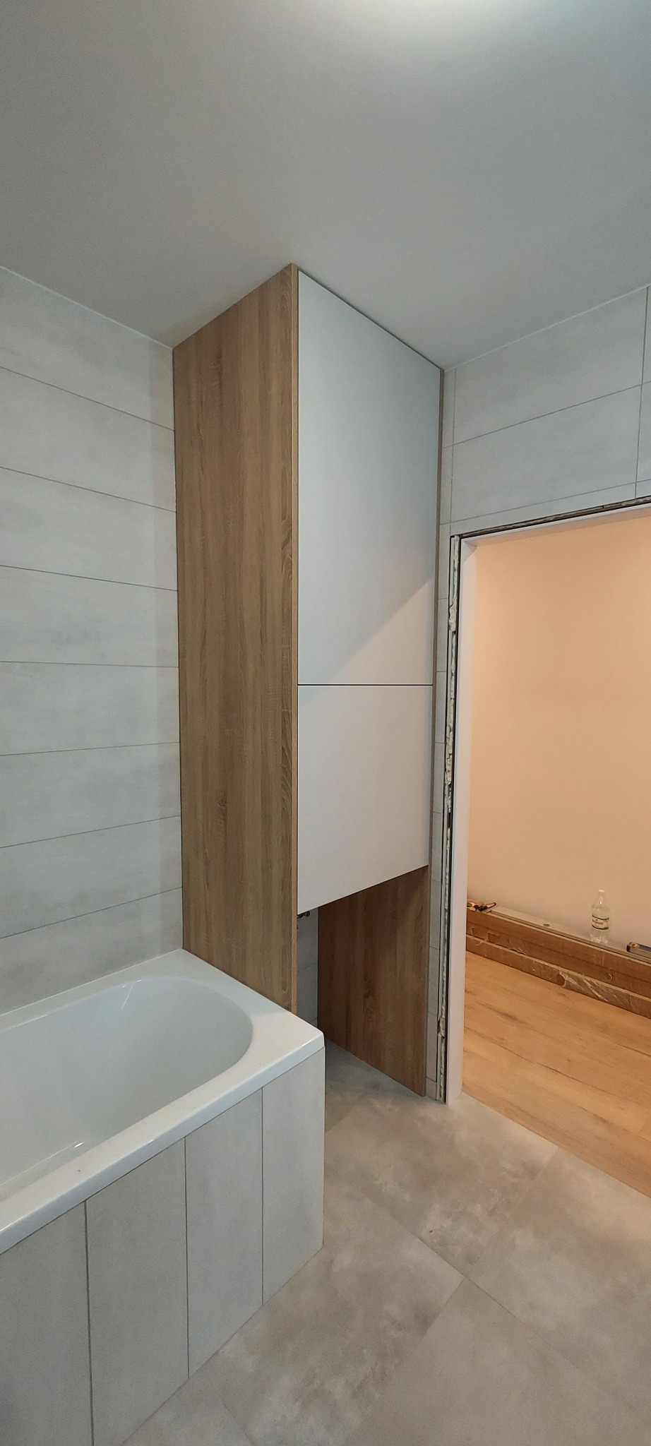 Półka łazienkowa z białymi frontami i bokami w kolorze jasnego drewna, zamontowana nad wanną w łazience z szarymi płytkami.