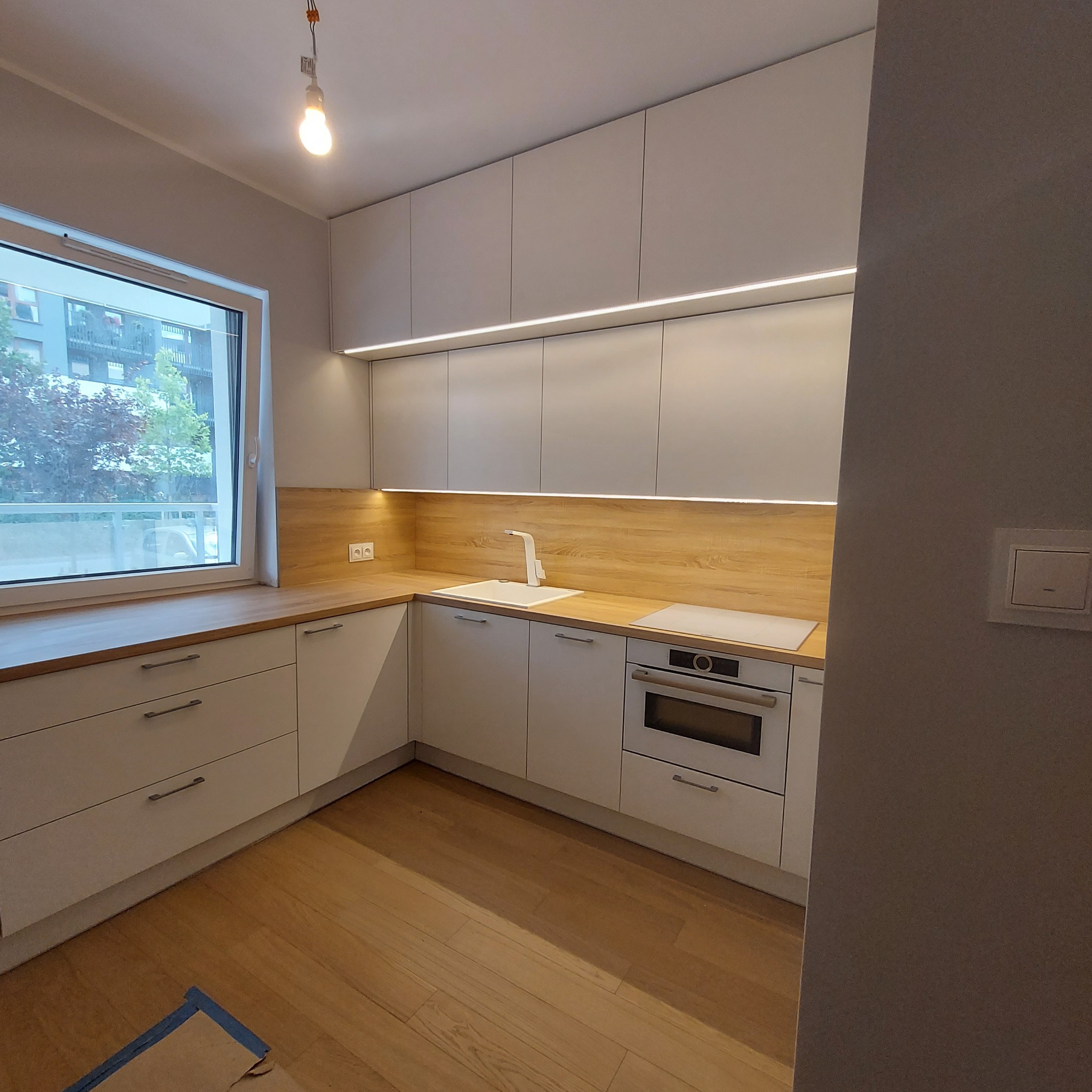 Jasna kuchnia z drewnianym blatem, oświetleniem LED pod szafkami górnymi i widokiem z okna na zewnątrz.