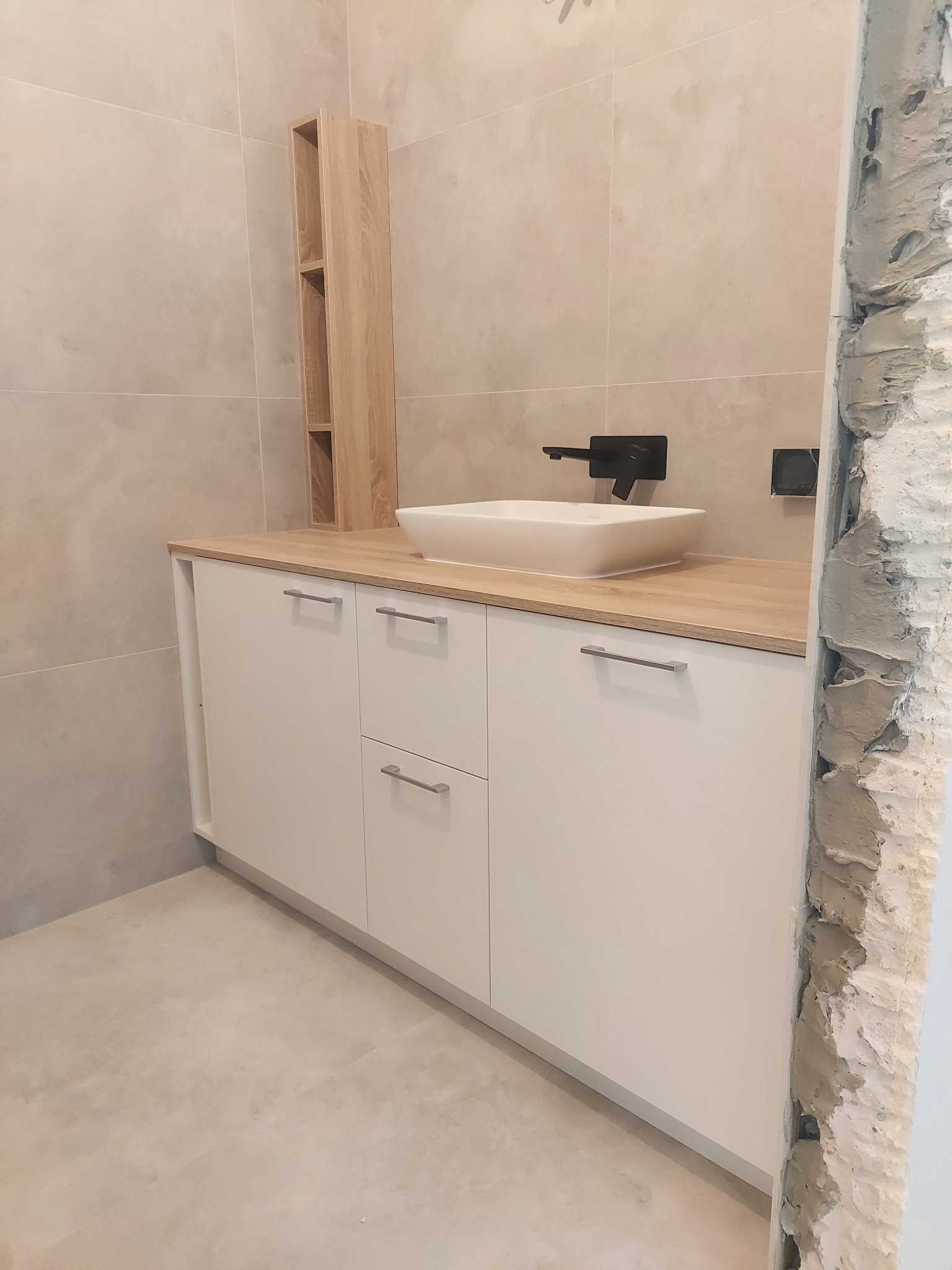 Nowoczesna łazienka w trakcie wykończenia, widoczna biała szafka pod umywalką z drewnianym blatem, wisząca półka z jasnego drewna, czarna bateria i fragment surowej ściany z widocznym tynkiem.