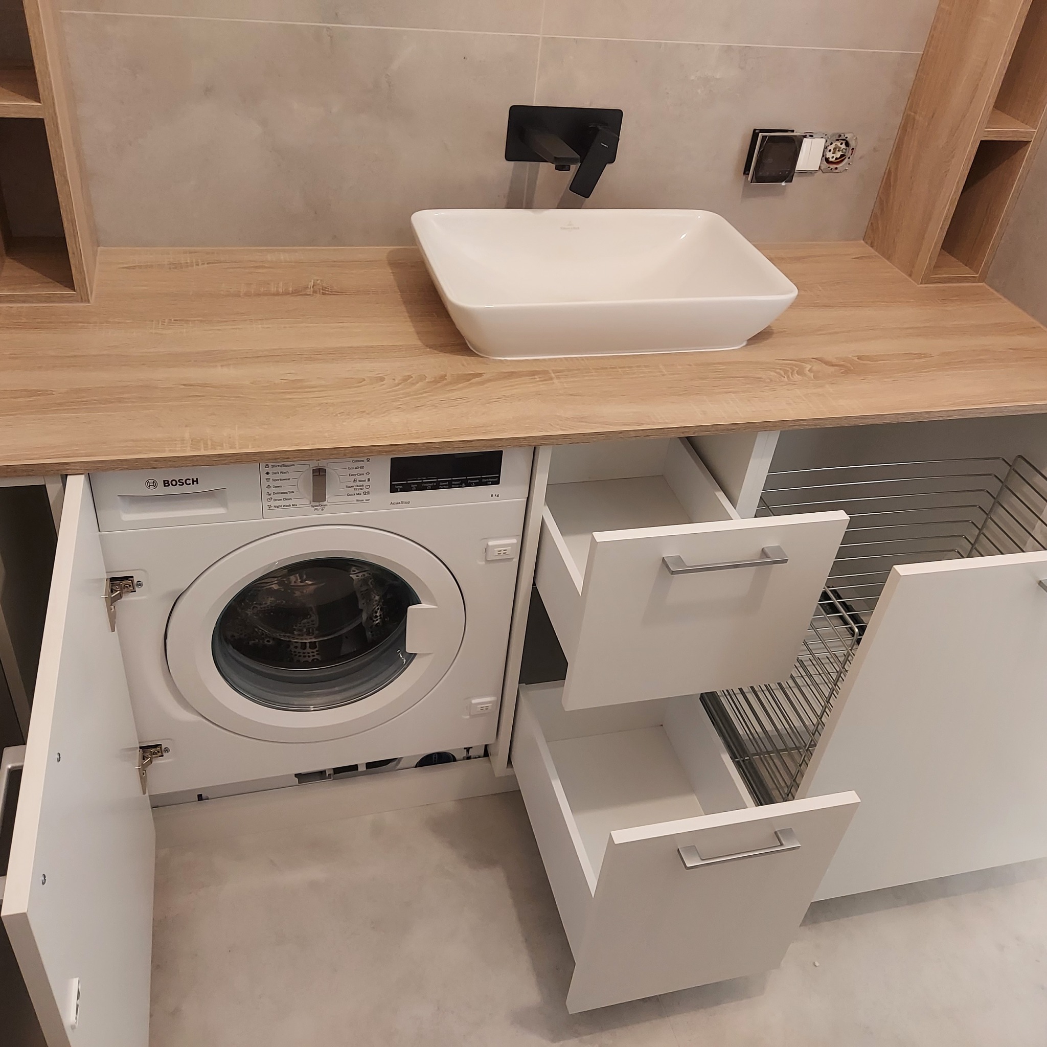 Łazienka z pralką Bosch ukrytą w szafce z blatem imitującym drewno, biała umywalka nablatowa i czarna bateria, otwarte białe szuflady i kosz na pranie.