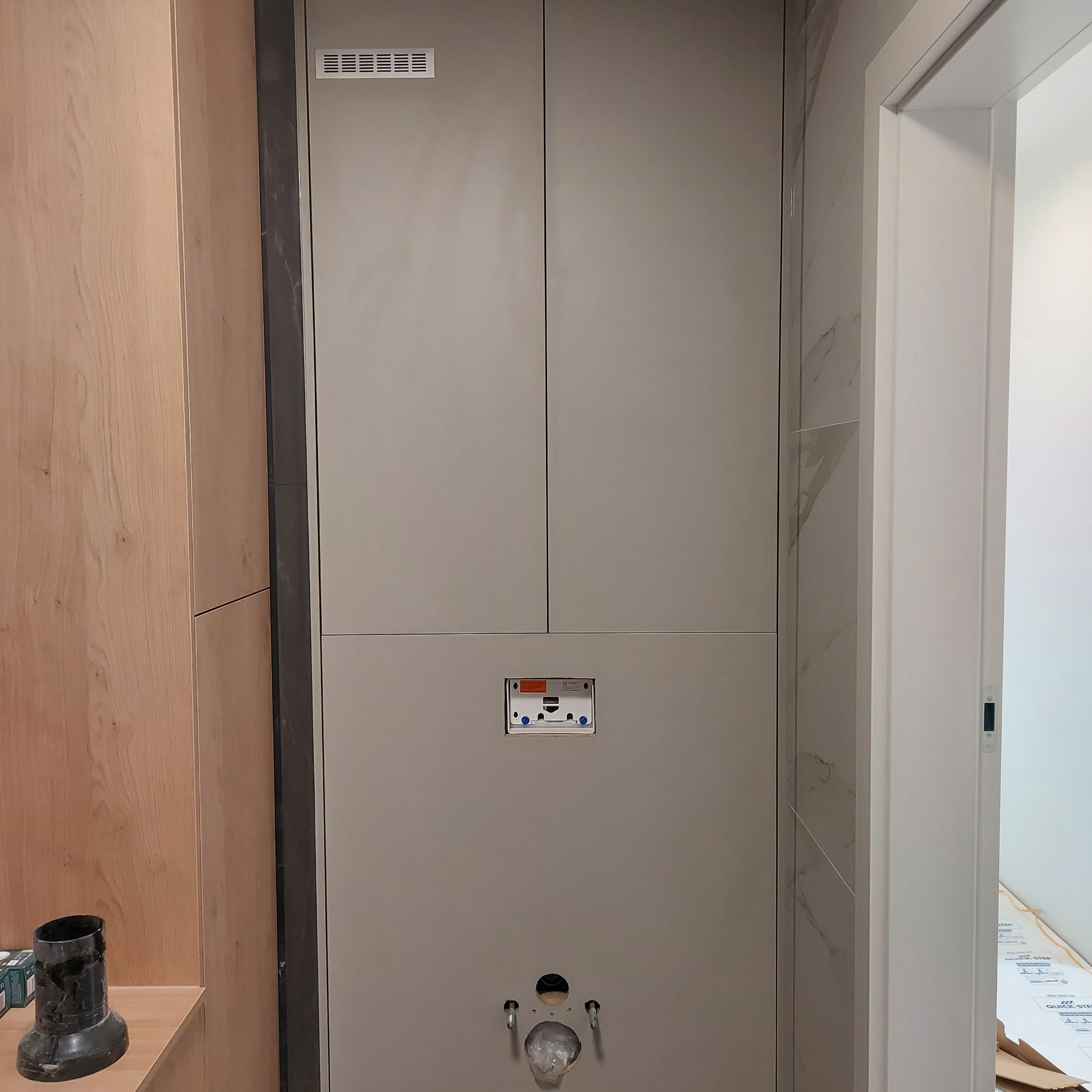 Montaż zabudowy WC ze spłuczką podtynkową i szafkami w łazience, widoczne przyłącza wodne i odpływowe, obok drewniana zabudowa i marmurowe wykończenie ściany.