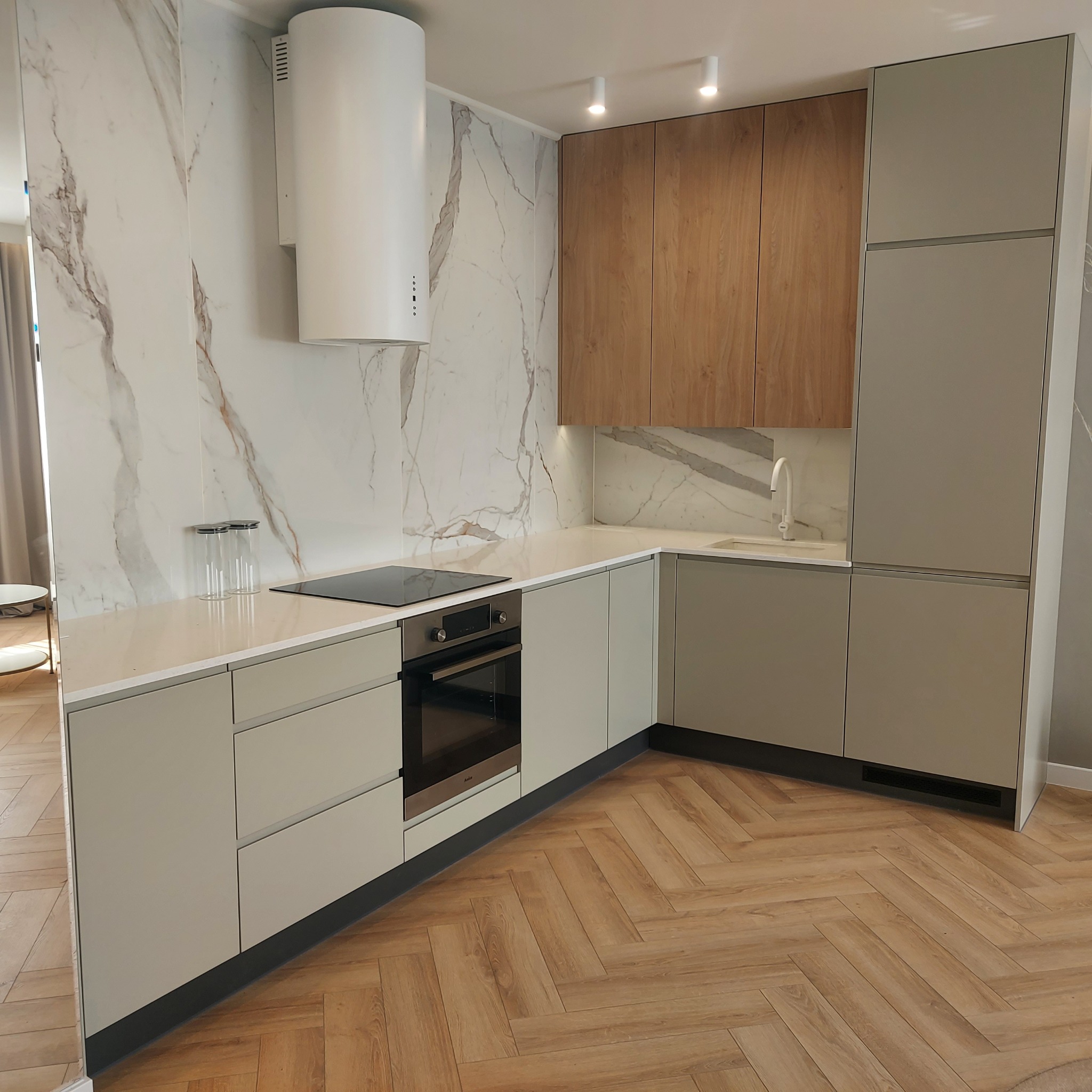 Nowoczesna kuchnia z szarymi i drewnianymi frontami, marmurowym panelem ściennym i podłogą ułożoną w jodełkę.