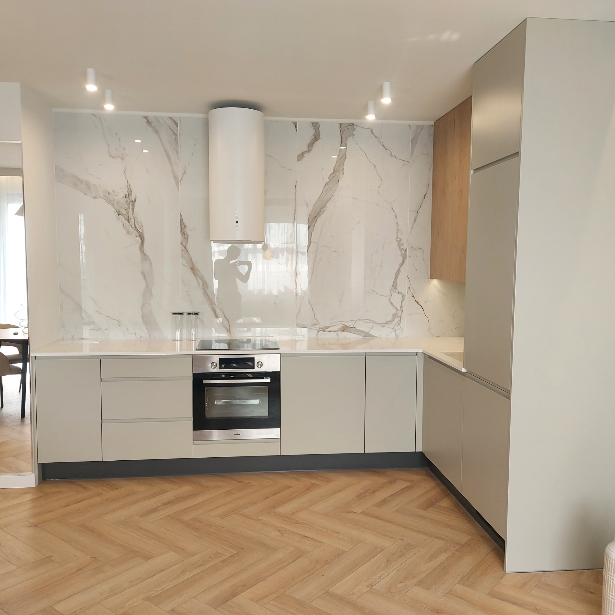 Nowoczesna kuchnia z jasnymi, gładkimi frontami szafek, marmurowym blatem i ścianą, drewnianymi akcentami i parkietem ułożonym w jodełkę.