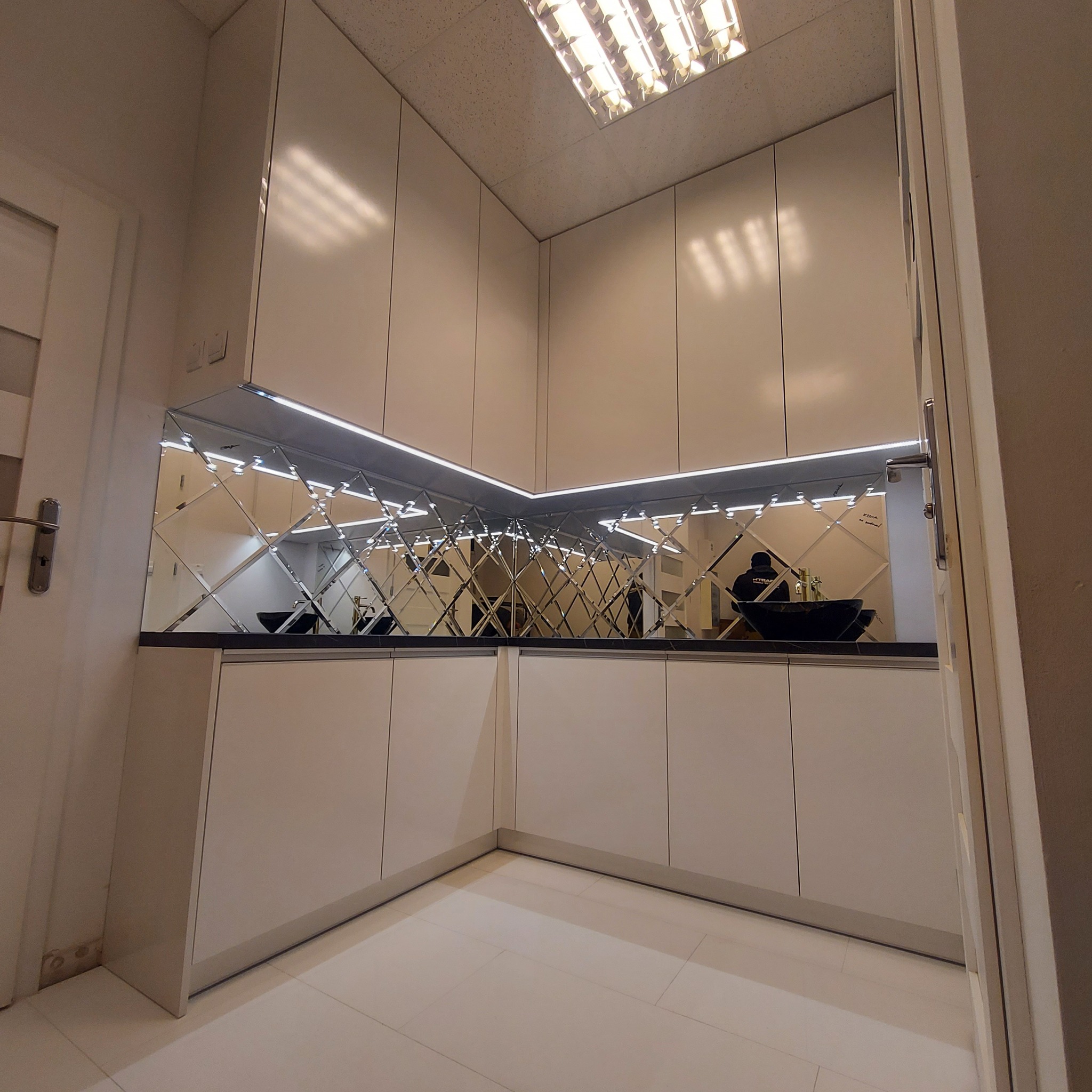 Nowoczesna kuchnia z białymi, lakierowanymi frontami, czarnym blatem i lustrzanym panelem pomiędzy szafkami górnymi i blatem, odbijającym sylwetkę człowieka.