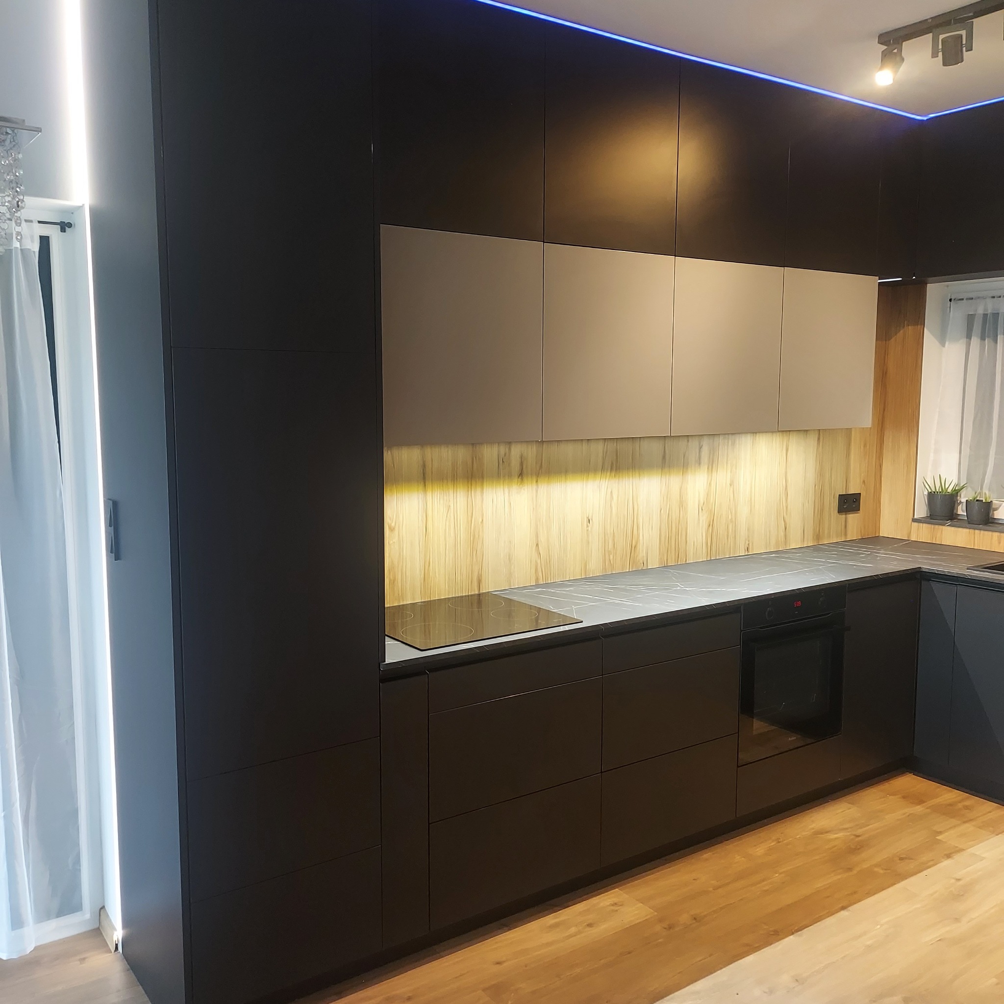 Nowoczesna kuchnia z czarnymi frontami szafek, jasnymi górnymi szafkami i drewnianym panelem ściennym podświetlonym od dołu, z blatem imitującym marmur i oświetleniem LED na suficie.