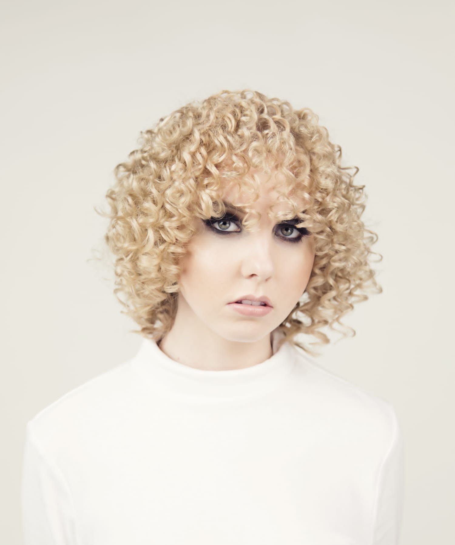 Portret kobiety z blond, kręconymi włosami w stylu afro i mocnym makijażem oczu, ubrana w biały golf.