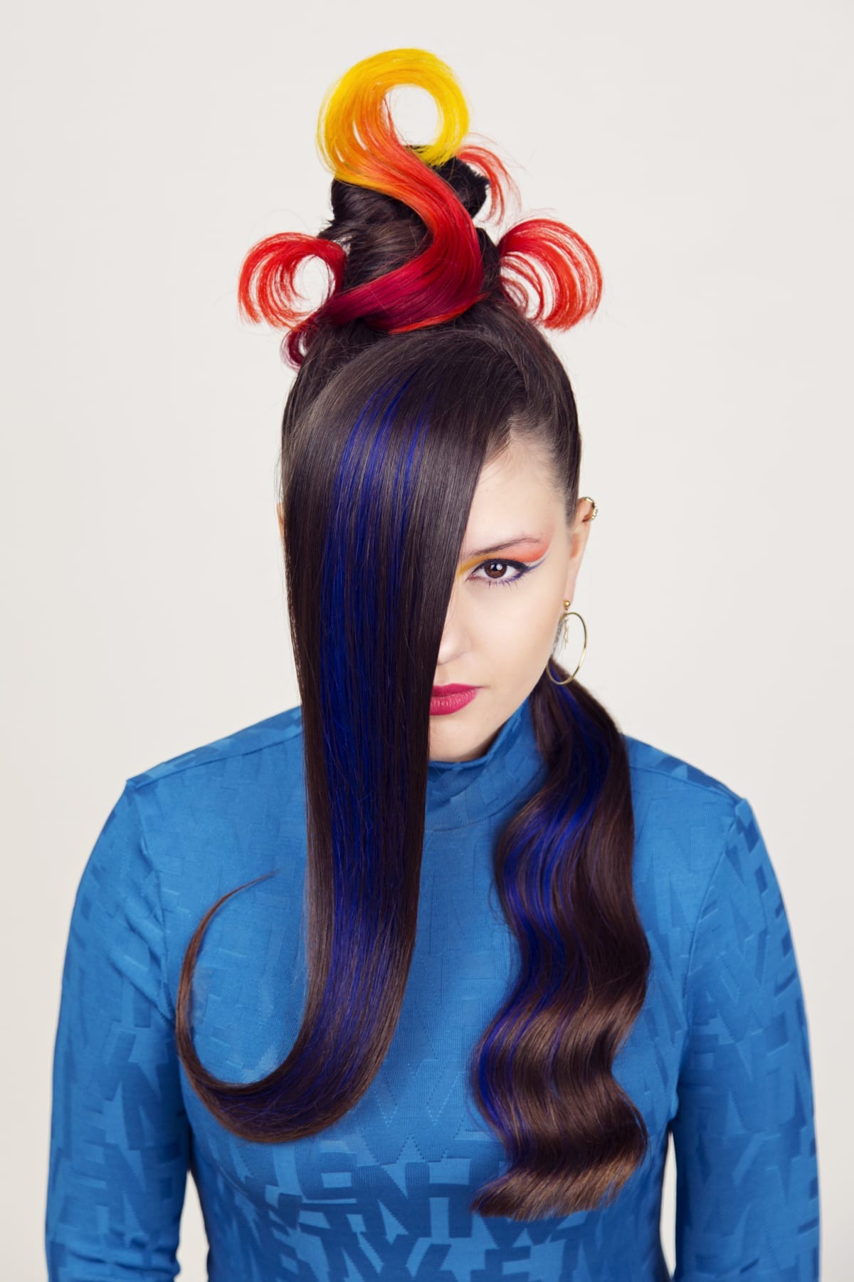 Awangardowa fryzura z asymetrycznym ułożeniem, pasemkami w kolorze niebieskim i upięciem z włosów w odcieniach żółci, czerwieni i pomarańczy, uzupełniona wyrazistym makijażem oczu i ust