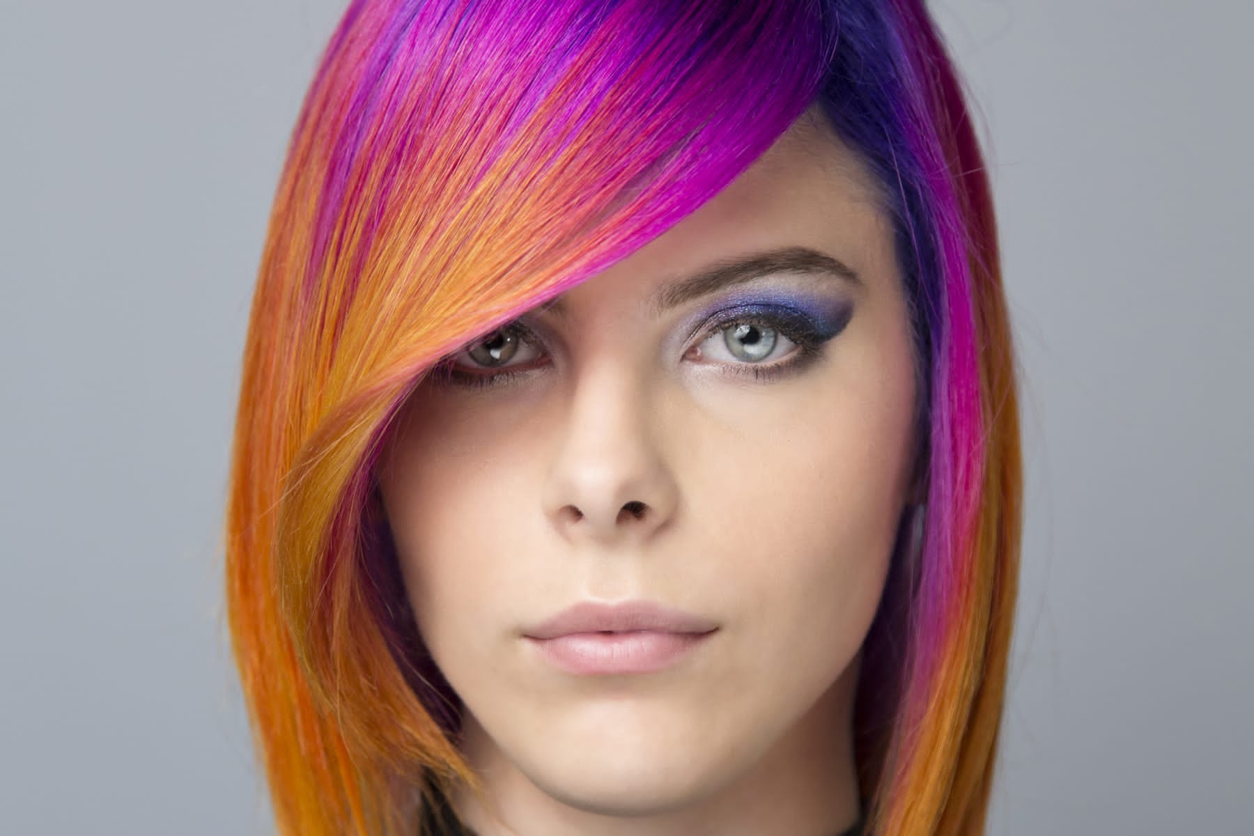 Portret kobiety z asymetryczną fryzurą, włosy farbowane w odcieniach fioletu, różu i pomarańczy, z intensywnym makijażem oczu w odcieniach granatu i fioletu.