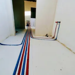 Instalacja wodna w bruzdach podłogi, rury czerwone i niebieskie prowadzą do ściany i rozdzielacza. Surowy stan wykończenia, widoczne niedoskonałości posadzki.