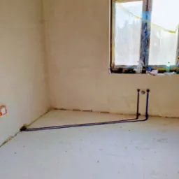 Wnętrze w stanie surowym z wyprowadzonymi rurami instalacji wodnej (czerwona i niebieska) przy ścianie z oknem zabezpieczonym folią malarską.