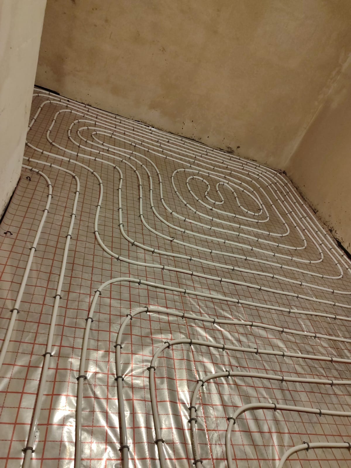 Białe rury ogrzewania podłogowego ułożone na folii z czerwoną siatką w pomieszczeniu o surowych ścianach.