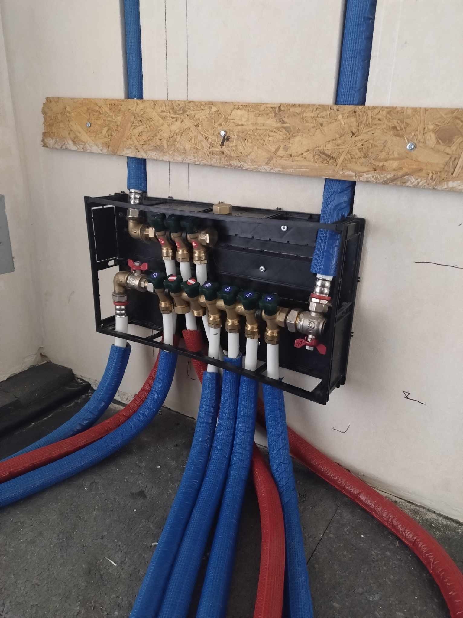 Rozdzielacz instalacji grzewczej z zaworami i rurami w izolacji niebieskiej i czerwonej, zamontowany na ścianie z surową płytą OSB powyżej.