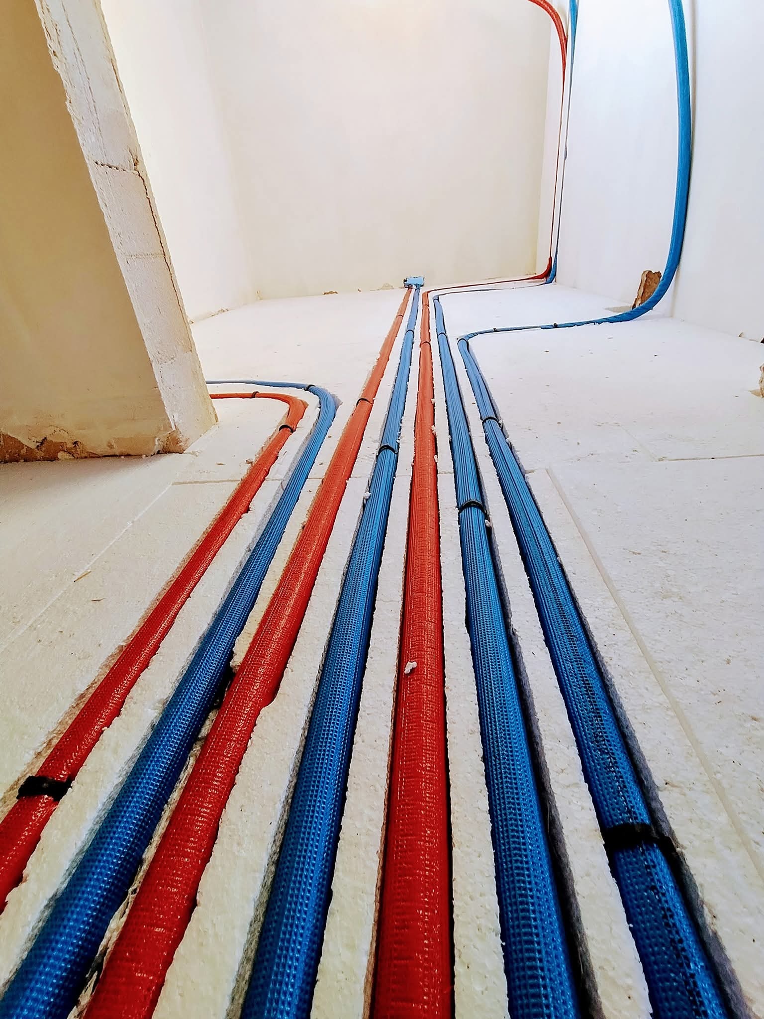 Podłogowe rury grzewcze w izolacji: czerwone i niebieskie, ułożone równolegle w białym styropianie, wnętrze w trakcie instalacji.