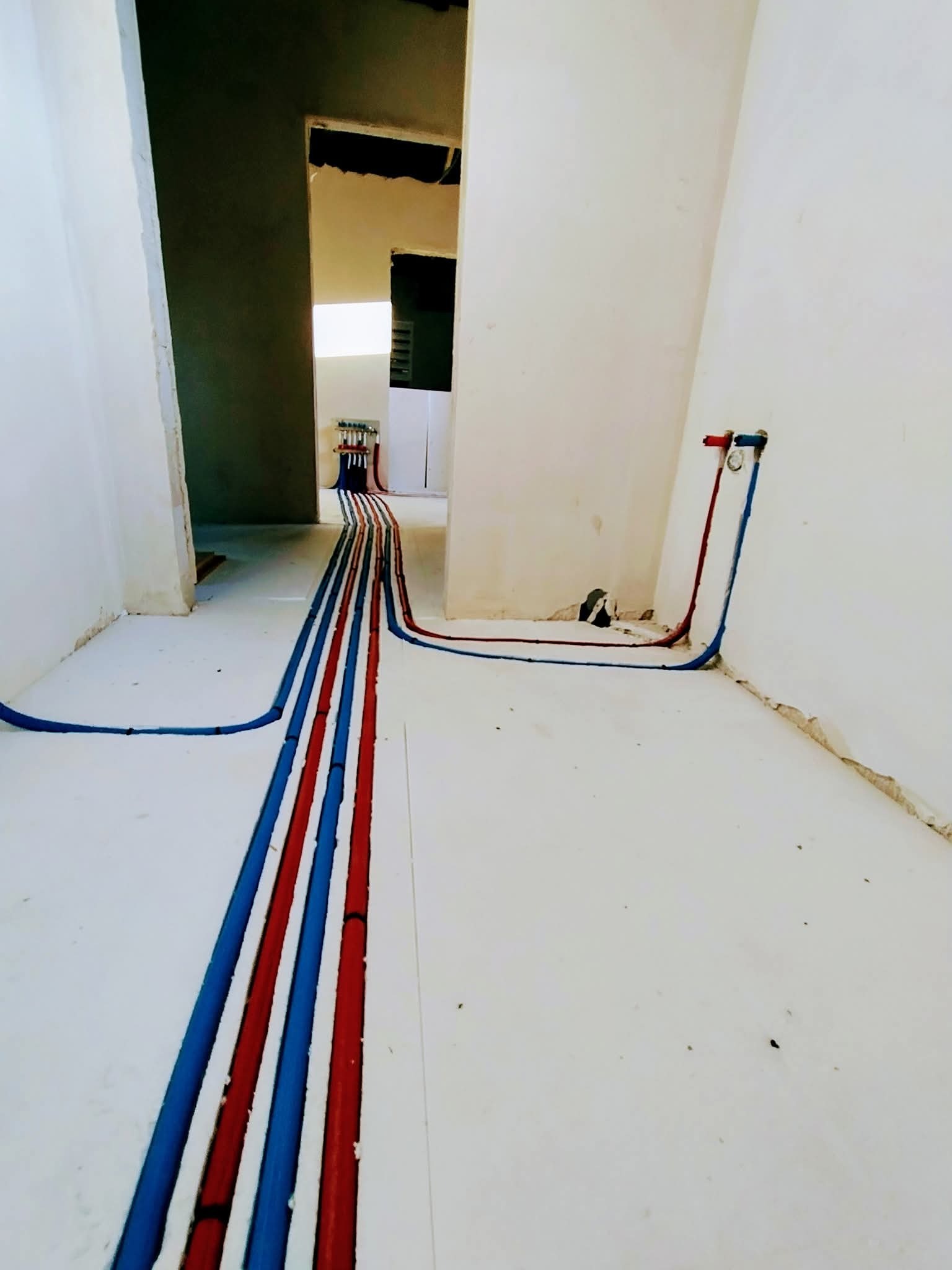 Instalacja wodna w bruzdach podłogi, rury czerwone i niebieskie prowadzą do ściany i rozdzielacza. Surowy stan wykończenia, widoczne niedoskonałości posadzki.