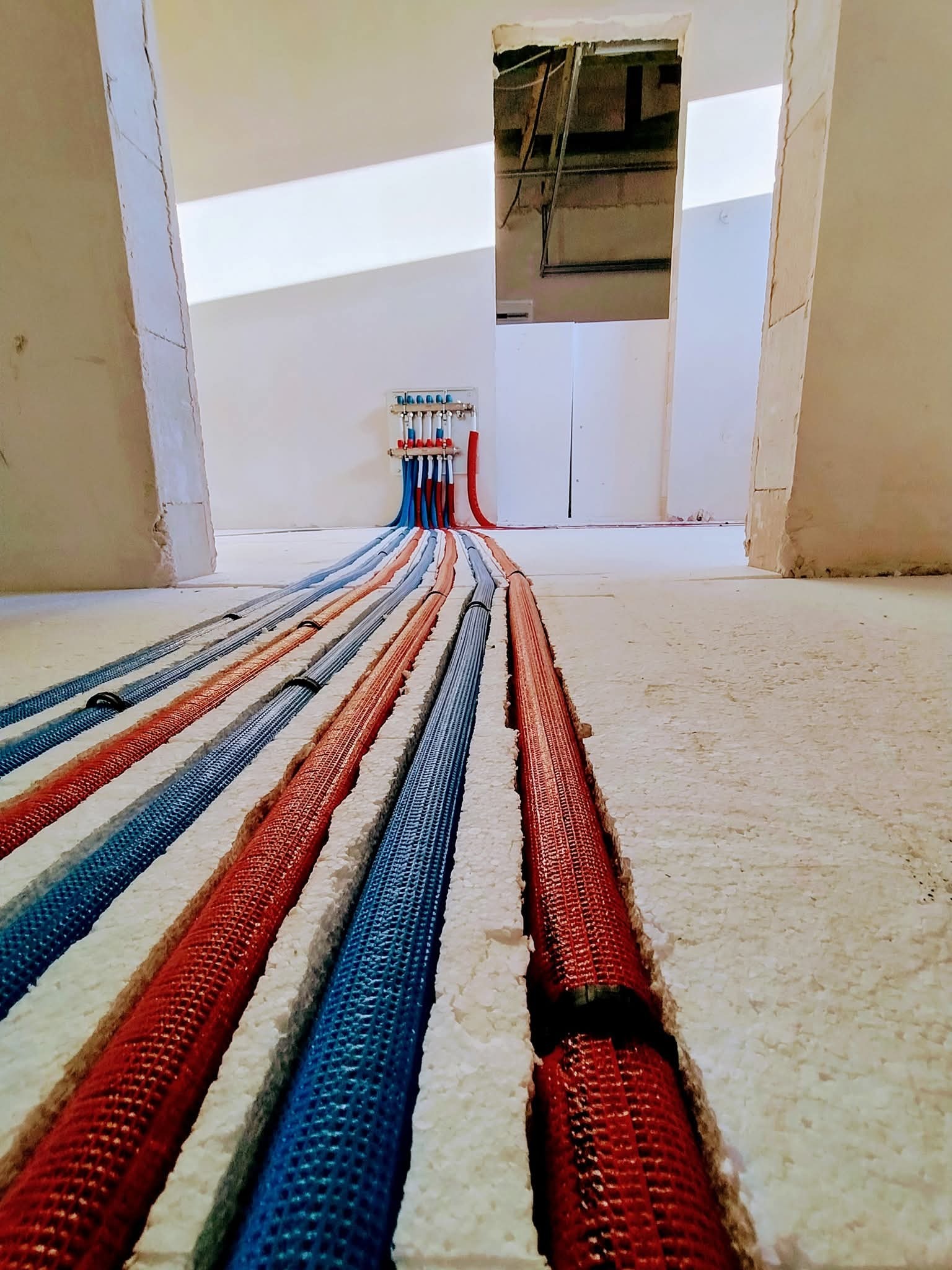 Podłogowe ogrzewanie centralne w izolacji: niebieskie i czerwone rury prowadzą do rozdzielacza w surowym wnętrzu. Widok z poziomu podłogi, w trakcie instalacji.