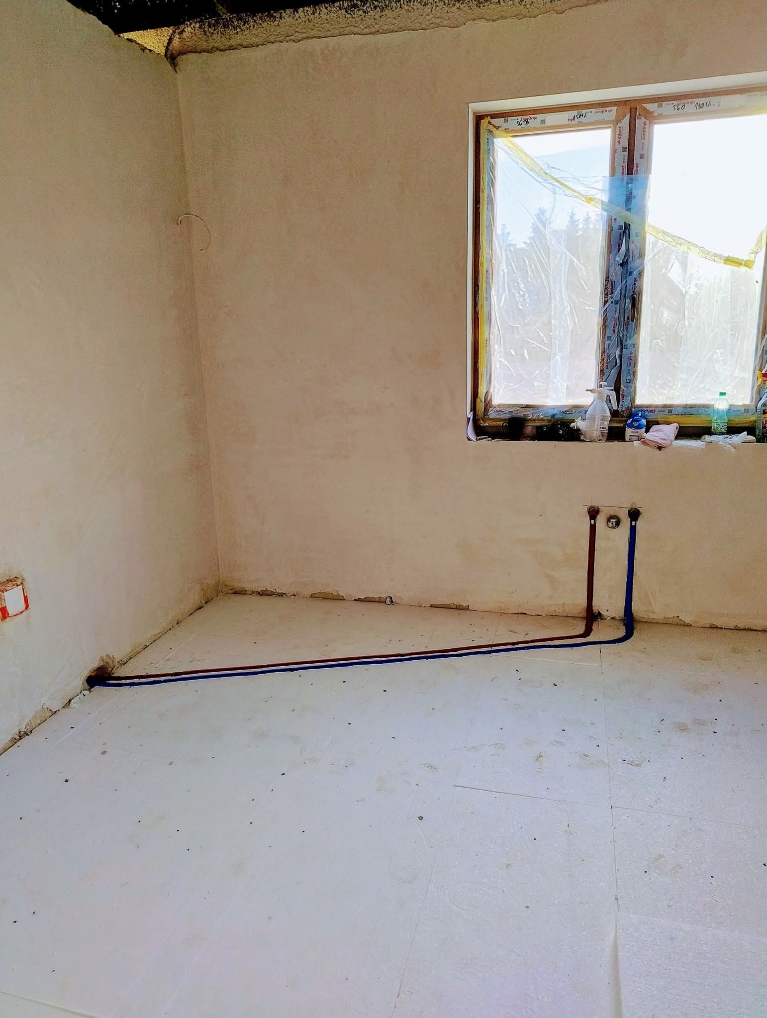 Wnętrze w stanie surowym z wyprowadzonymi rurami instalacji wodnej (czerwona i niebieska) przy ścianie z oknem zabezpieczonym folią malarską.