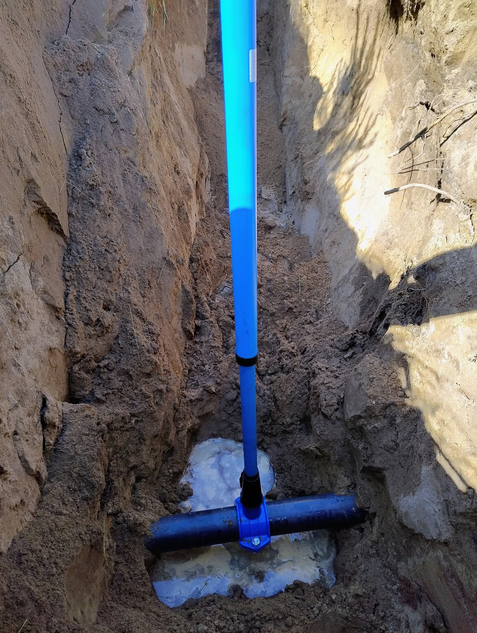 Niebieski zacisk na czarnej rurze w wykopie w Więcborku, prawdopodobnie świeżo zamontowany. Woda w wykopie. Ściany wykopu z ziemi piaszczystej.