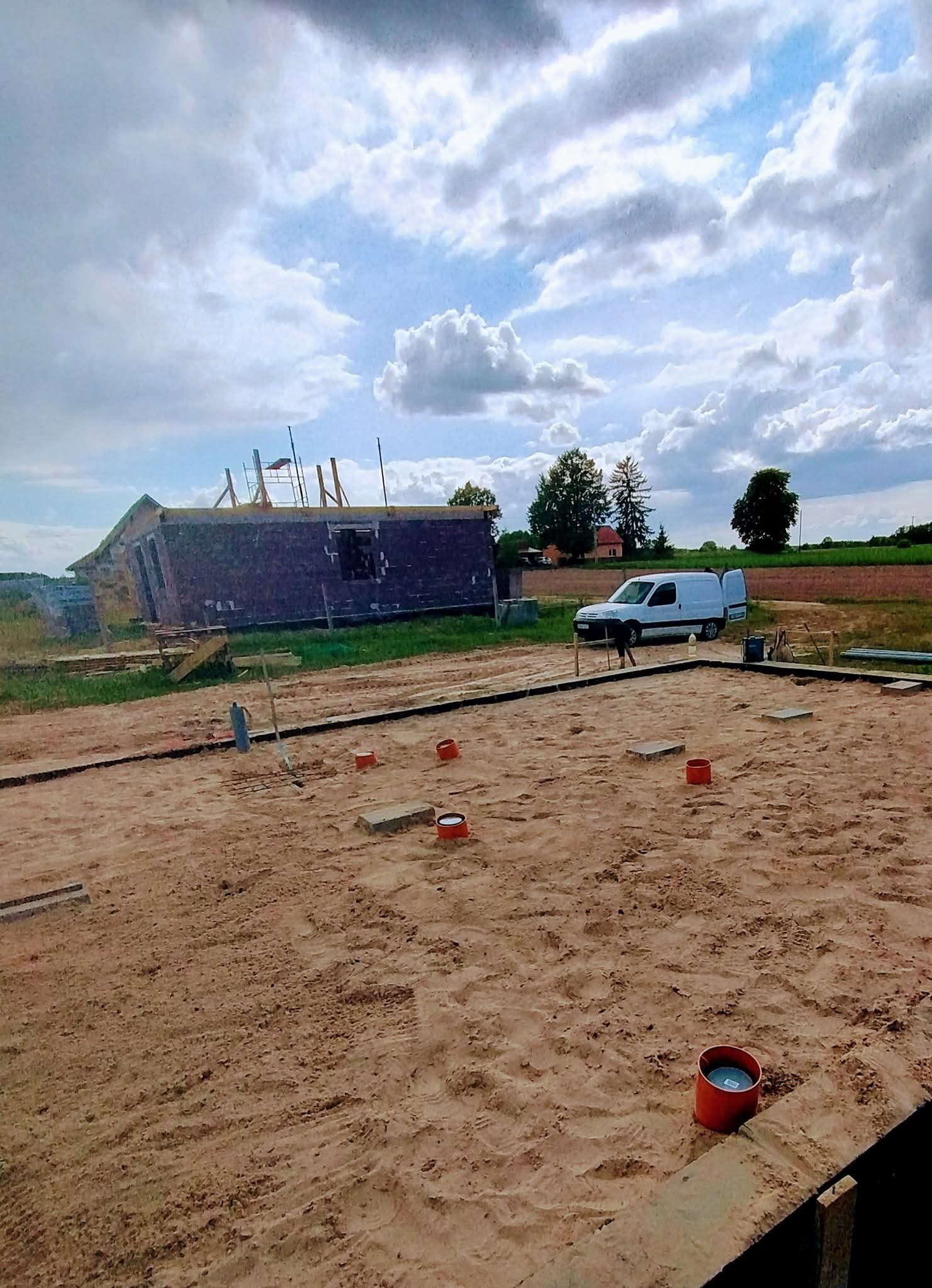 Fundamenty domu w trakcie budowy, widoczne rury instalacji wodno-kanalizacyjnej, na drugim planie budynek w stanie surowym otwartym i biały bus.
