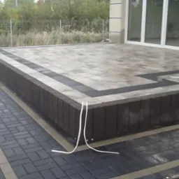 Ułożona kostka brukowa w odcieniach szarości i czerni tworzy taras z obrzeżem. Widoczne białe kable i budynek w tle. Geometryczny wzór i precyzja wykonania.