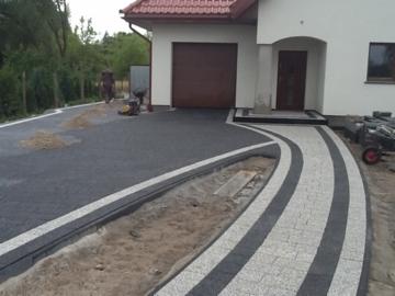 Nowoczesny podjazd z kostki brukowej w odcieniach szarości i bieli, z łagodnym łukiem prowadzącym do domu z brązowym dachem i garażem.