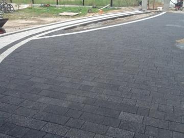 Ułożona ciemnoszara kostka brukowa z białym pasem dekoracyjnym, tworząca ścieżkę i podjazd. Widoczne elementy krajobrazu i fragment wózka budowlanego w tle.