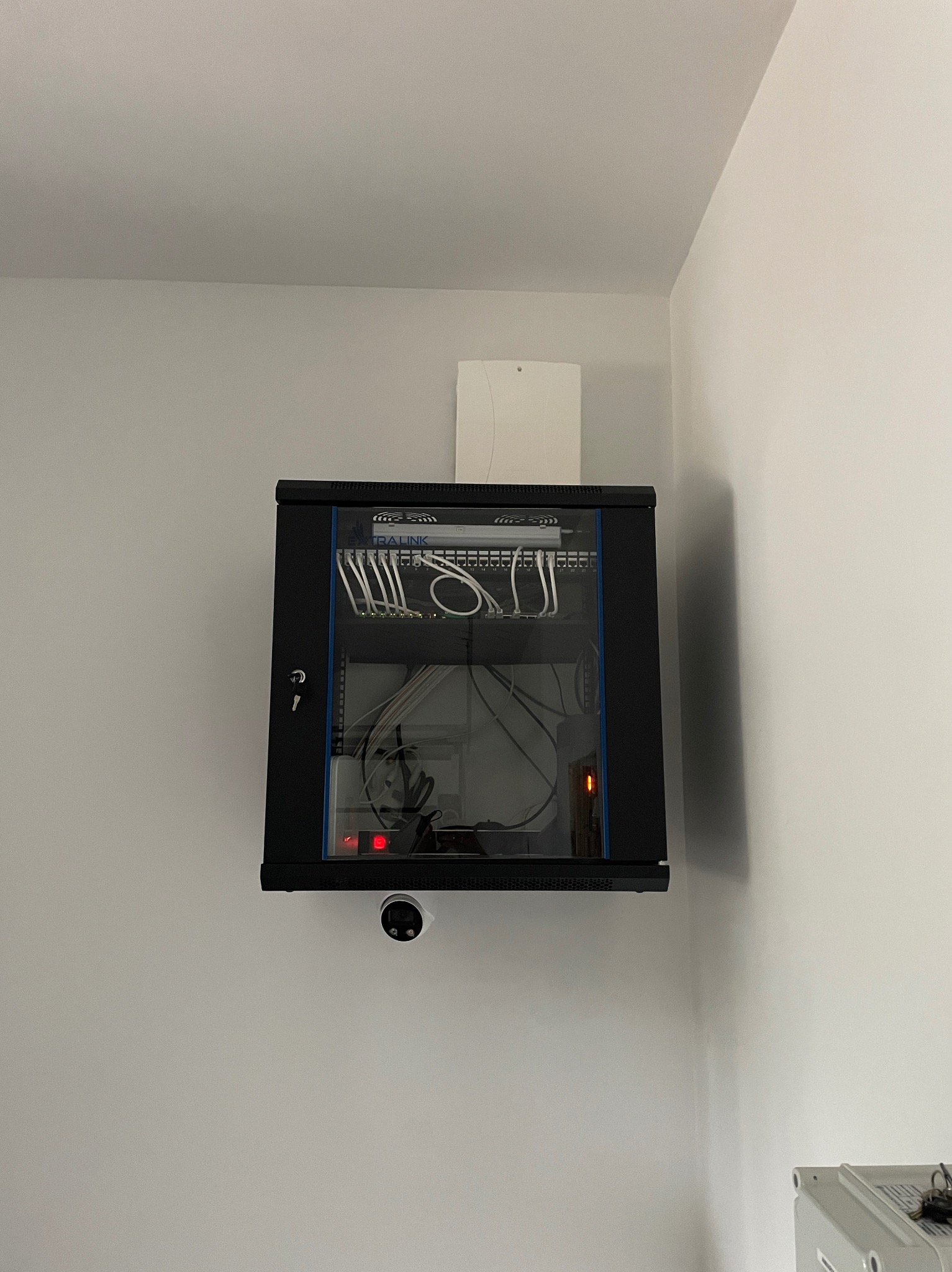 Zamknięta szafa rack z okablowaniem sieciowym, zamontowana na ścianie, pod nią widoczna kamera monitoringu.