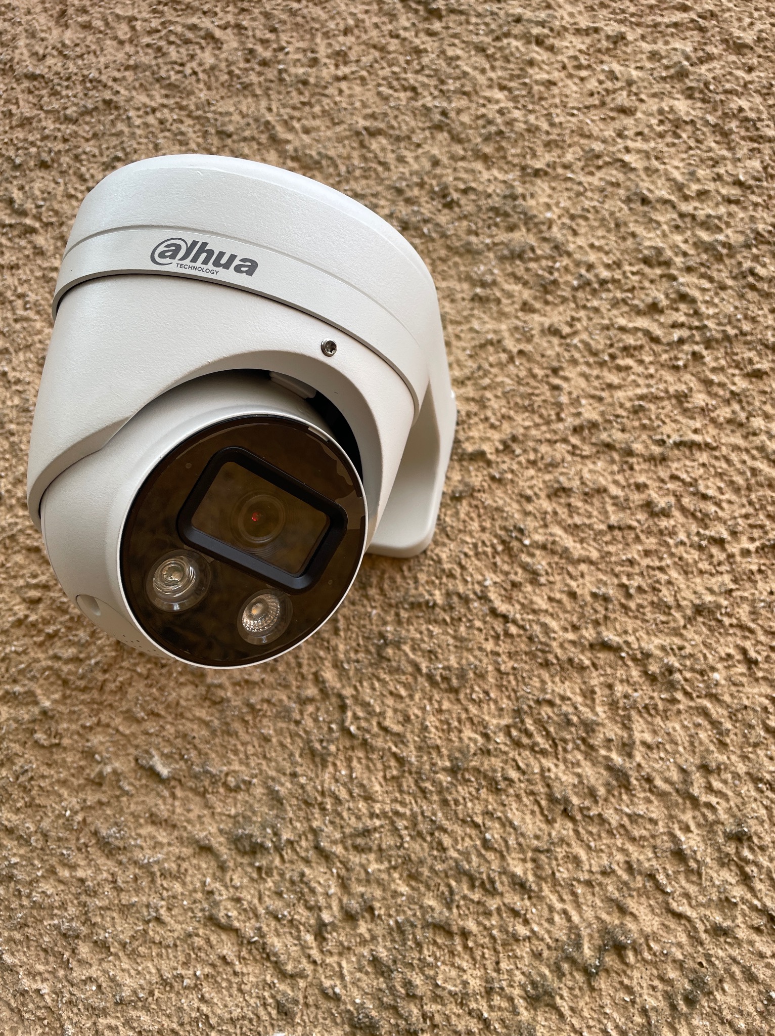 Kamera Dahua na teksturowanej, beżowej ścianie. Widoczne logo producenta i diody IR. Ujęcie z dołu, podkreślające solidność montażu. Dyskretny element systemu zabezpieczeń.