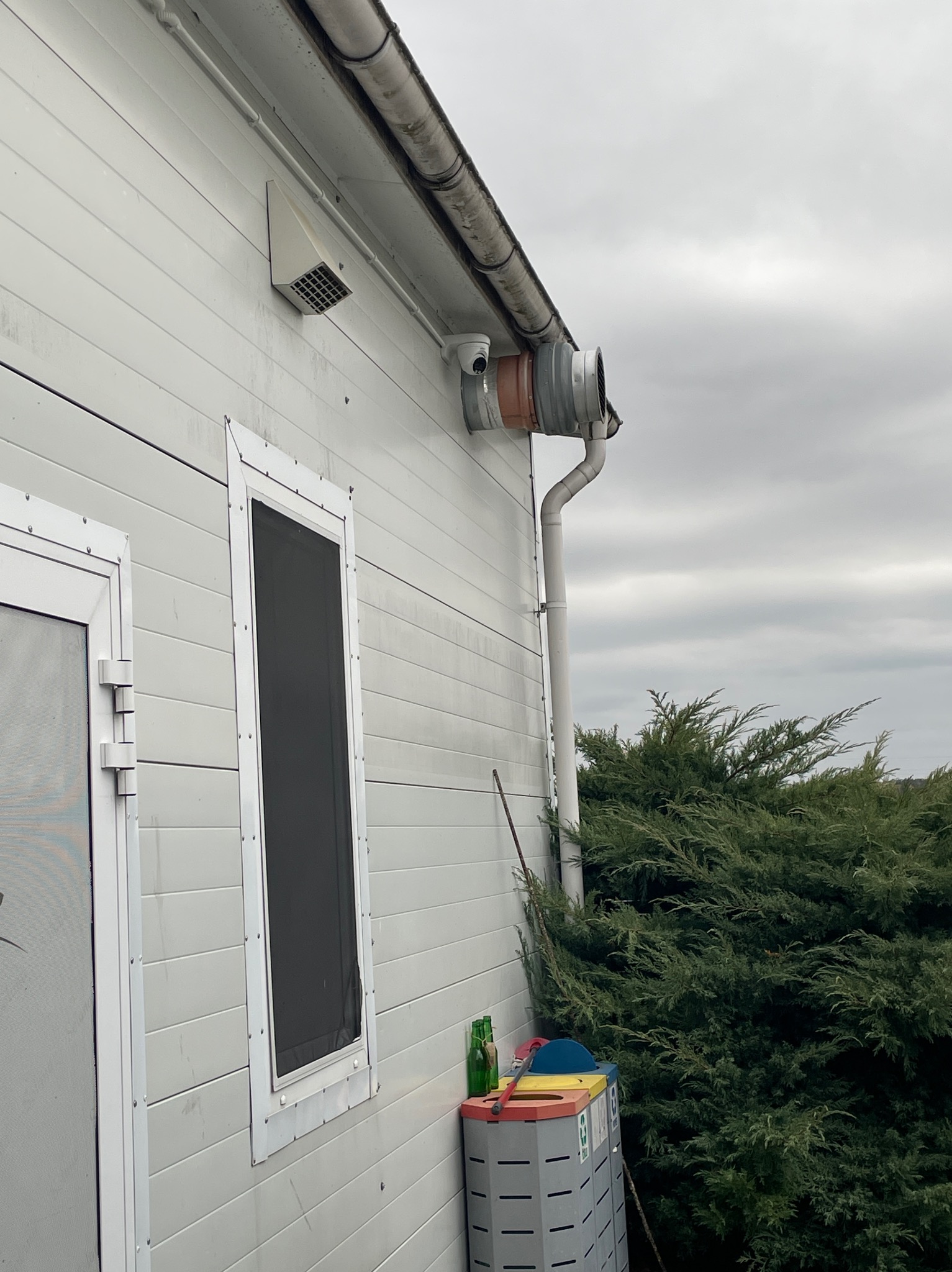 Kamera monitoringu zamontowana na elewacji budynku, obok rury spustowej i wentylacji. Widoczny fragment okna z siatką i kosz na śmieci. Pochmurne niebo w tle.