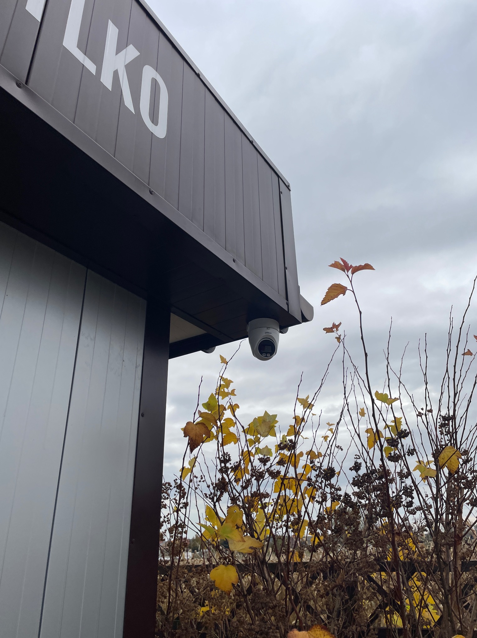 Kamera kopułkowa zamontowana pod zadaszeniem budynku z napisem 'LKO', widoczna na tle pochmurnego nieba i krzewów z żółtymi liśćmi.