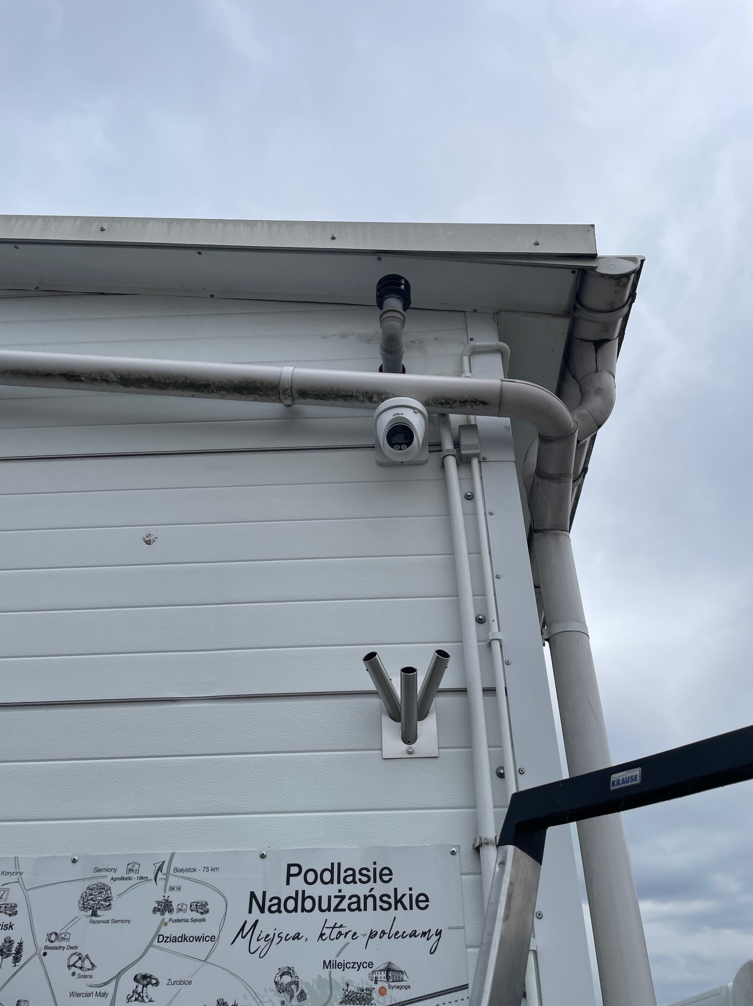 Kamera monitoringu Dahua zamontowana na białej ścianie budynku z rurami i tablicą informacyjną. Widok z dołu, pochmurne niebo w tle.