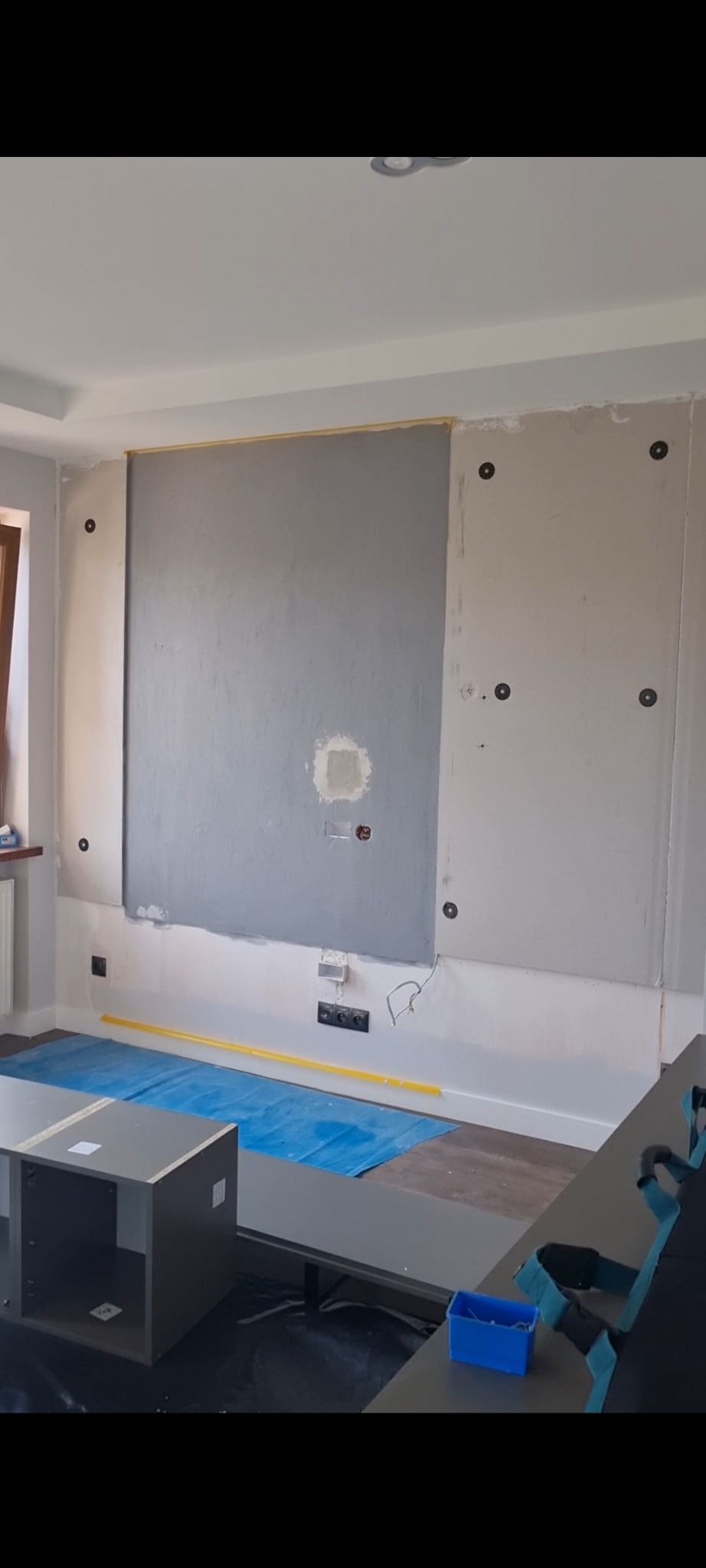 Ściana w trakcie przygotowania do montażu telewizora, widoczne otwory na kable, listwa przypodłogowa zabezpieczona folią i taśmą malarską, obok skrzynka z narzędziami.