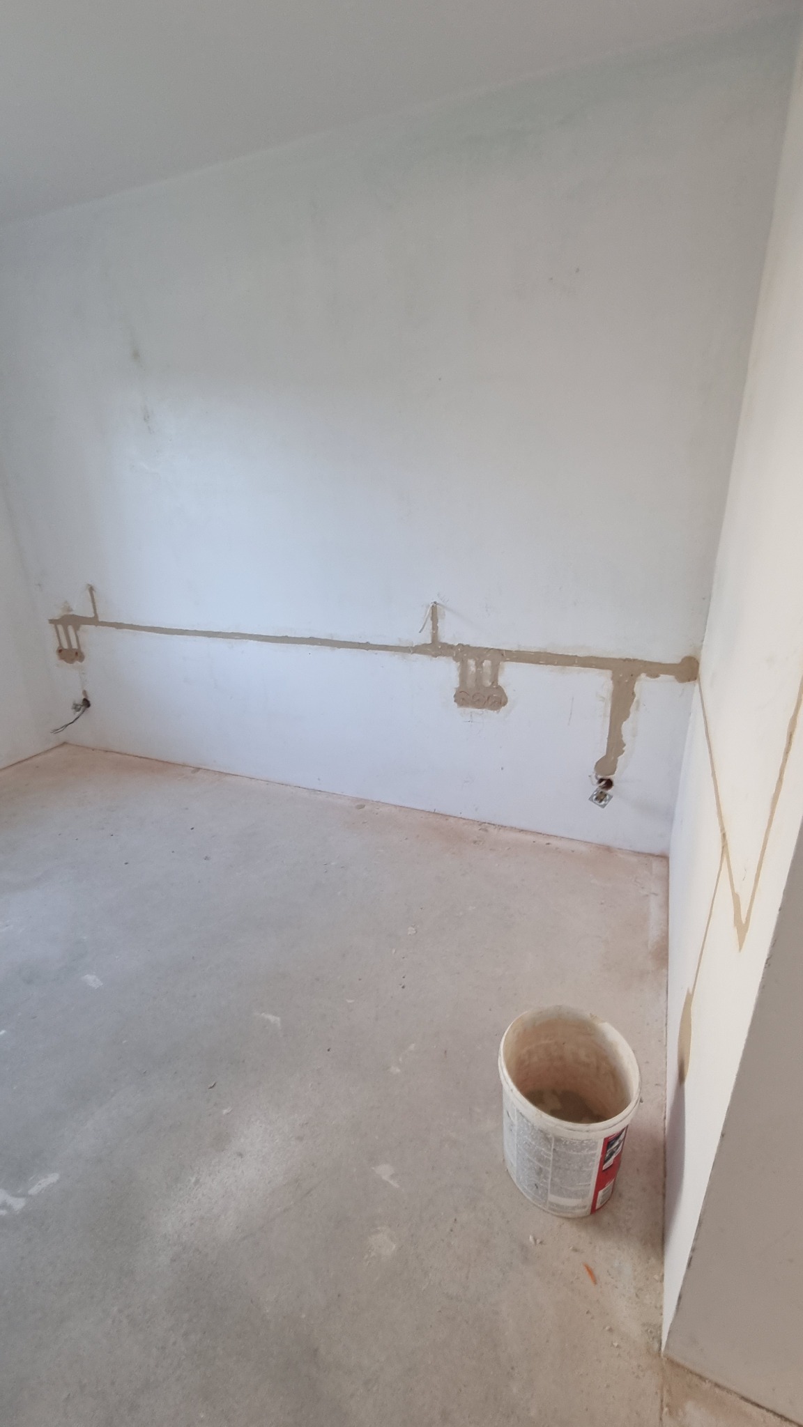 Wnętrze w trakcie prac instalacyjnych: widoczne bruzdy w ścianie z wyprowadzonymi przewodami elektrycznymi, puszki instalacyjne zamurowane, na podłodze wiadro po farbie.