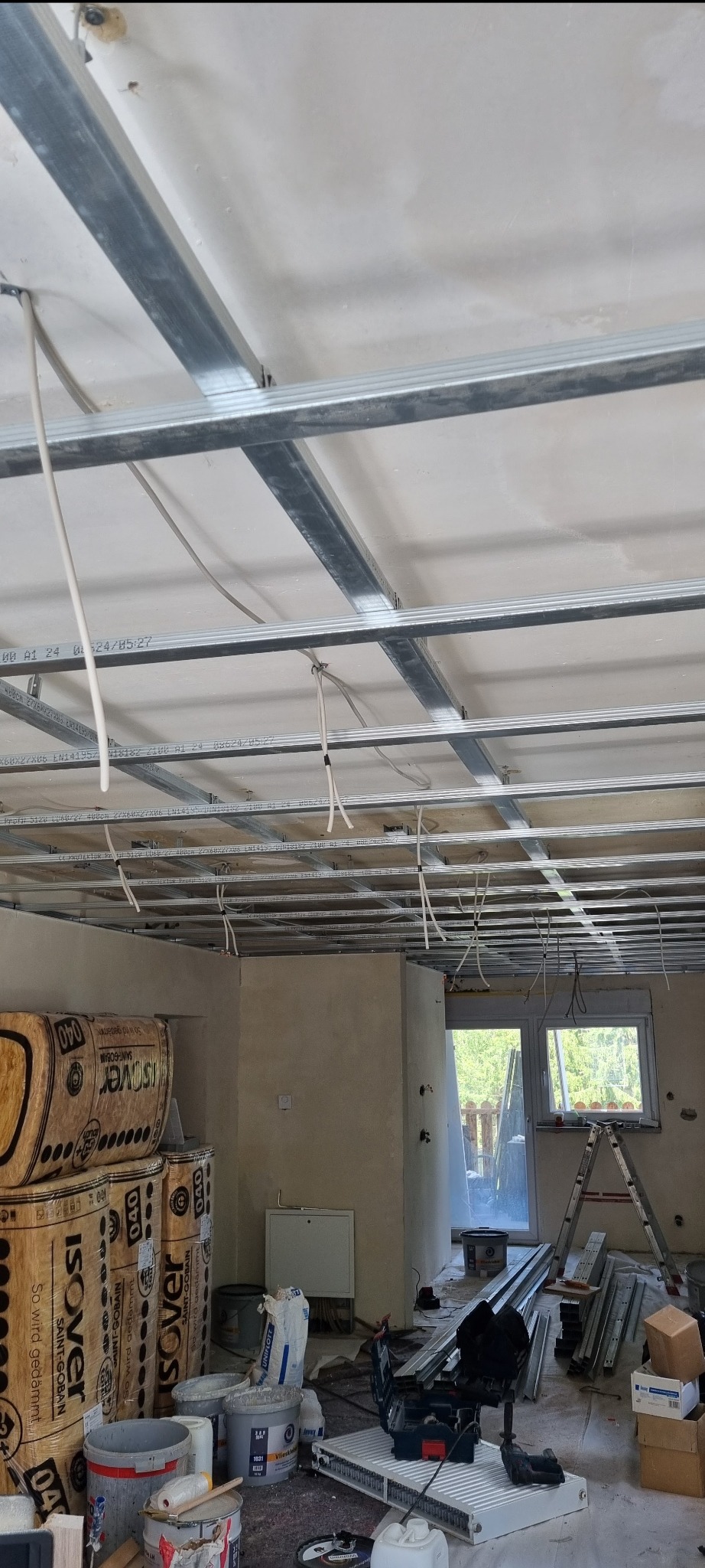 Montaż sufitu podwieszanego z widoczną konstrukcją metalową, wystającymi przewodami elektrycznymi i stosem izolacji Isover na podłodze w trakcie remontu.