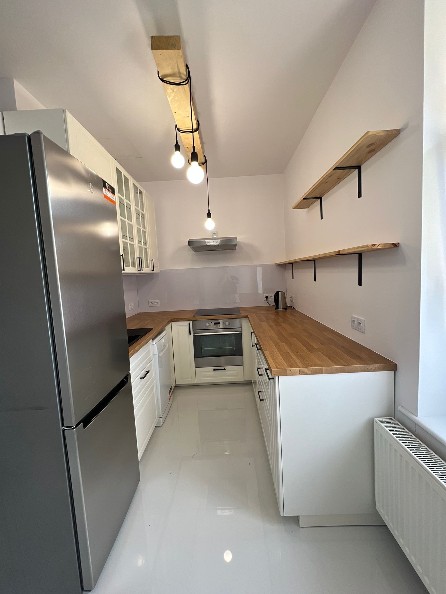 Nowoczesna, jasna kuchnia z białymi szafkami i drewnianym blatem, oświetlona trzema żarówkami zwisającymi z drewnianej belki. Na ścianie widoczne drewniane półki na czarnych wspornikach...