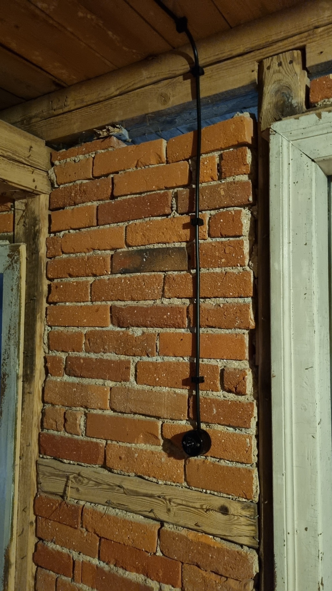 Czarny kabel elektryczny poprowadzony po ceglanej ścianie w starym budynku, przymocowany uchwytami, zakończony okrągłą, czarną puszką elektryczną, widoczne drewniane elementy konstrukcyjne...