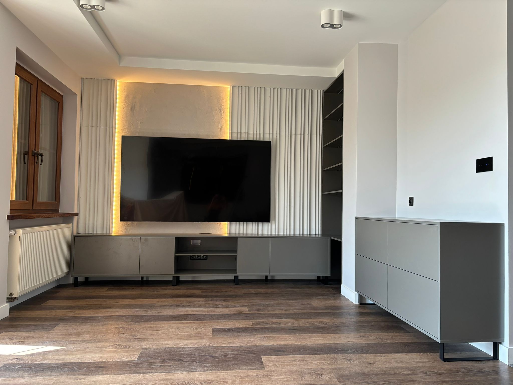 Nowoczesny salon z podświetlaną ścianą TV, szarą szafką RTV na nóżkach, drewnianą podłogą i grzejnikiem pod oknem.