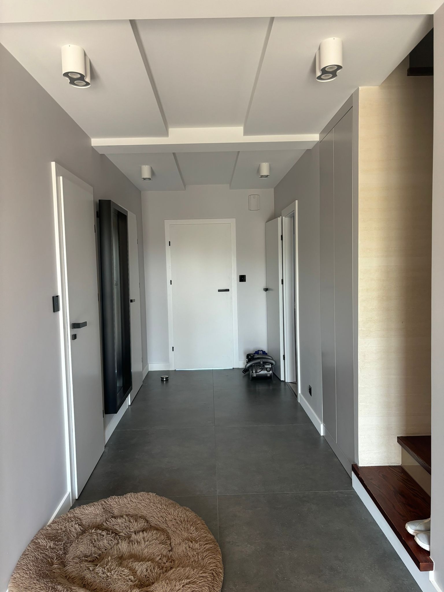 Nowoczesny przedpokój z szarą podłogą, białymi ścianami, wbudowanymi szafami i okrągłymi lampami sufitowymi z podwójnym oświetleniem, widoczny odkurzacz i legowisko dla zwierząt.