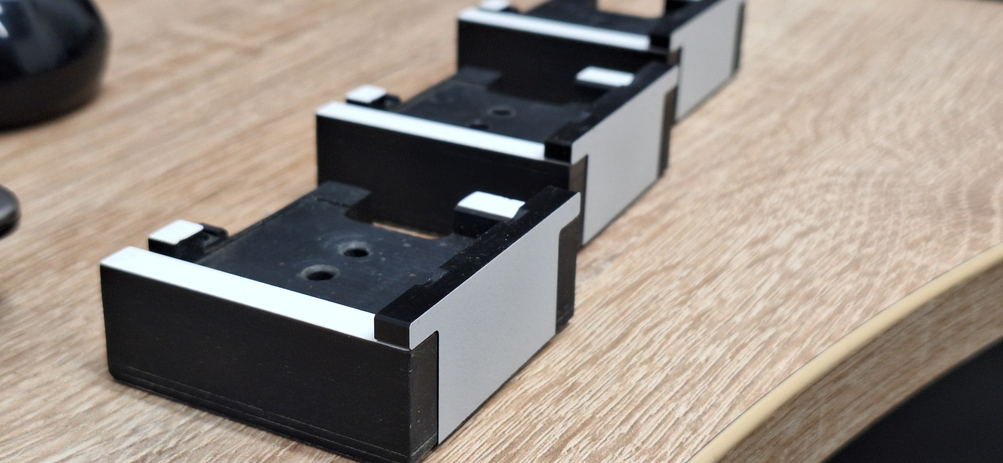 Trzy czarno-szare, identyczne elementy z tworzywa sztucznego ułożone jeden za drugim na drewnianej powierzchni, widoczne otwory montażowe.