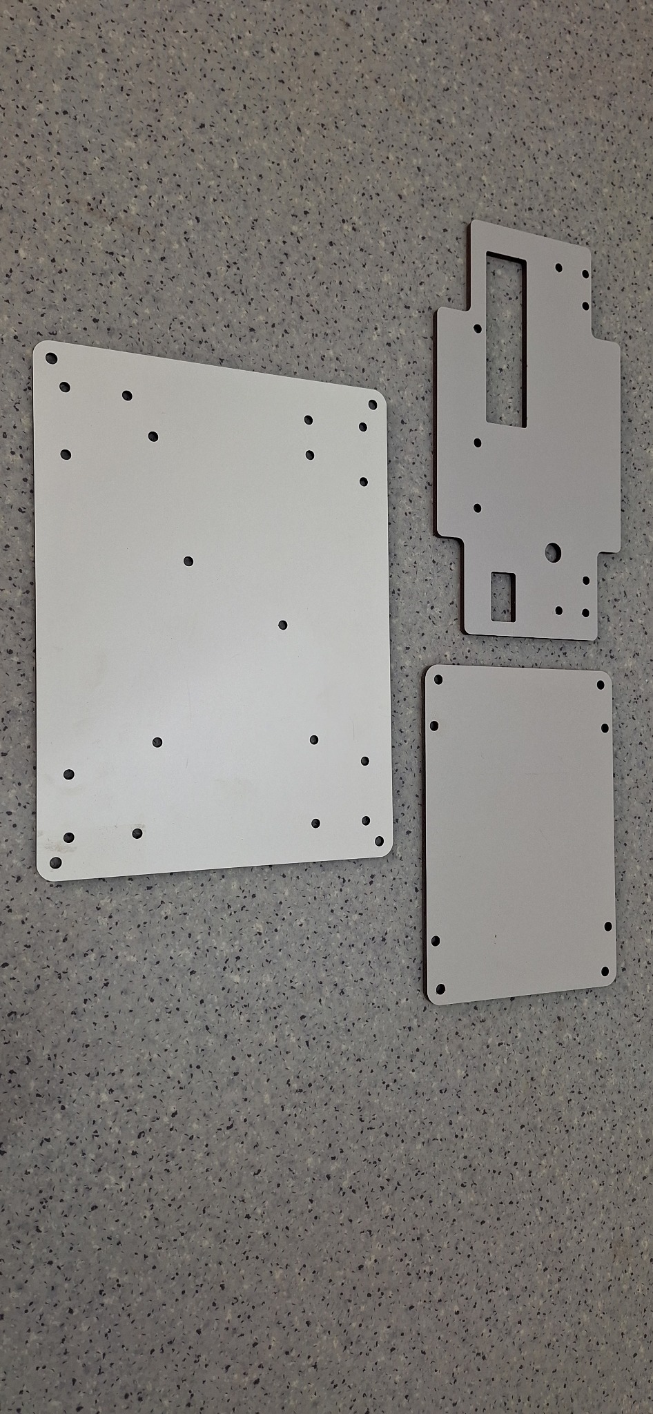 Trzy precyzyjnie wycięte, metalowe płyty z otworami, leżące na szarej, nakrapianej powierzchni.