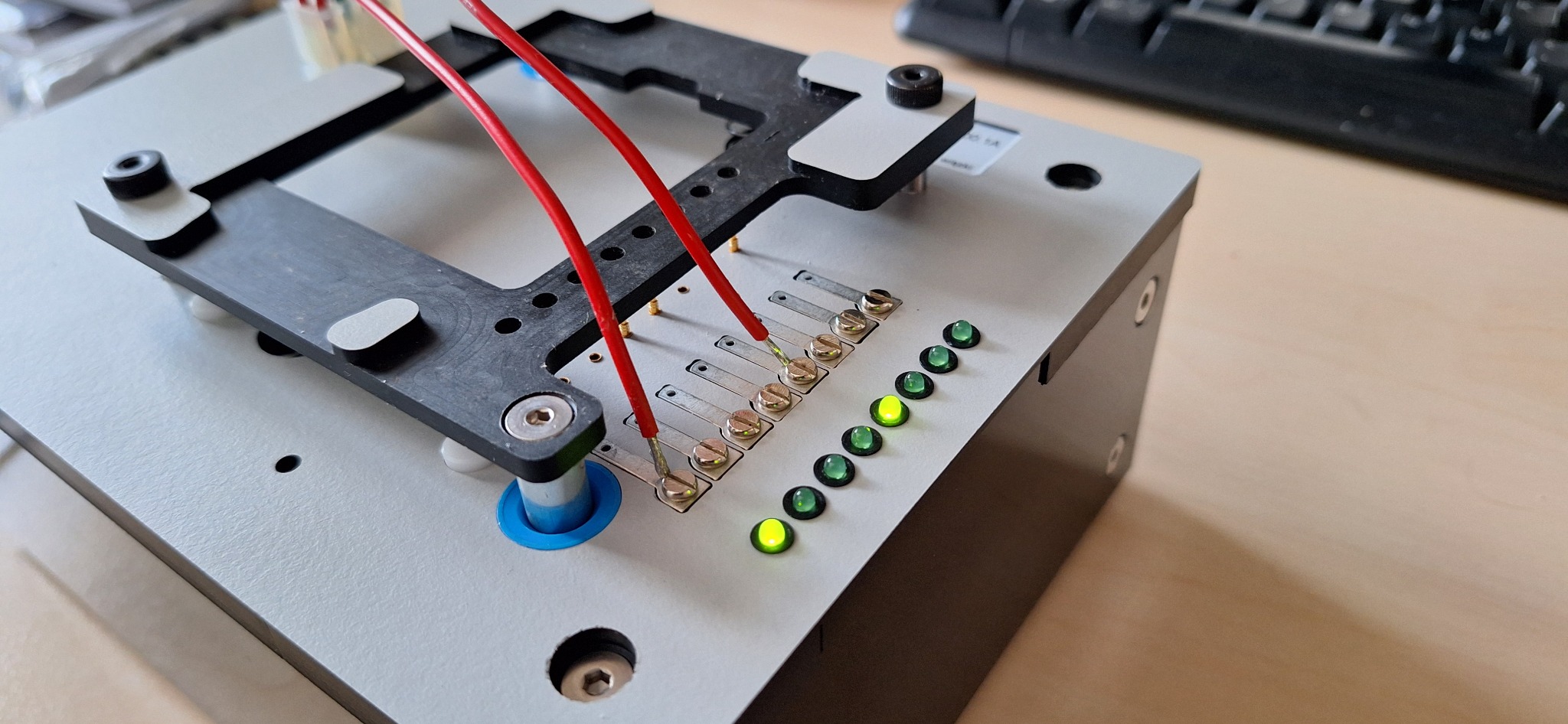 Metalowa platforma testowa z przewodami, śrubami i diodami LED, częściowo widoczna klawiatura w tle.