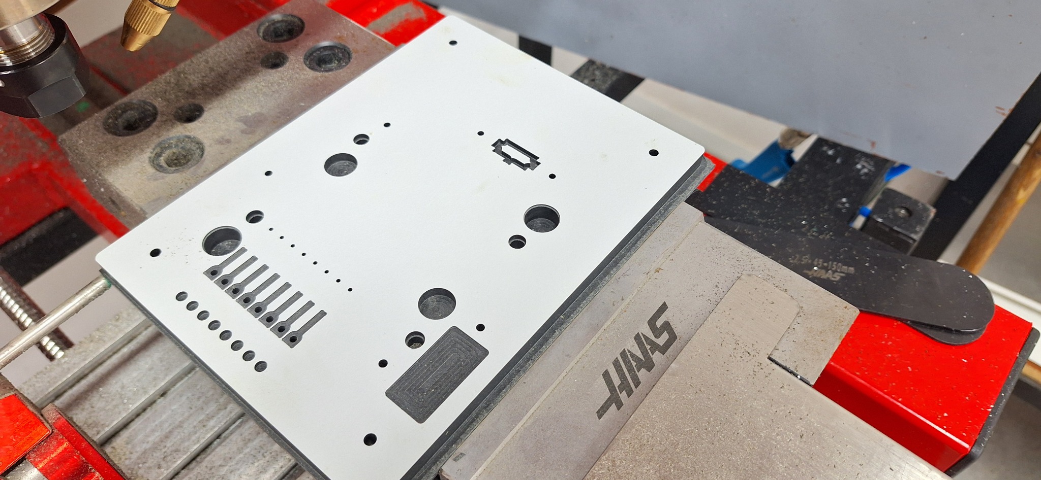 Biała płyta z wyfrezowanymi otworami i rowkami, zamocowana w uchwycie maszyny Haas, widoczna głowica frezarki.