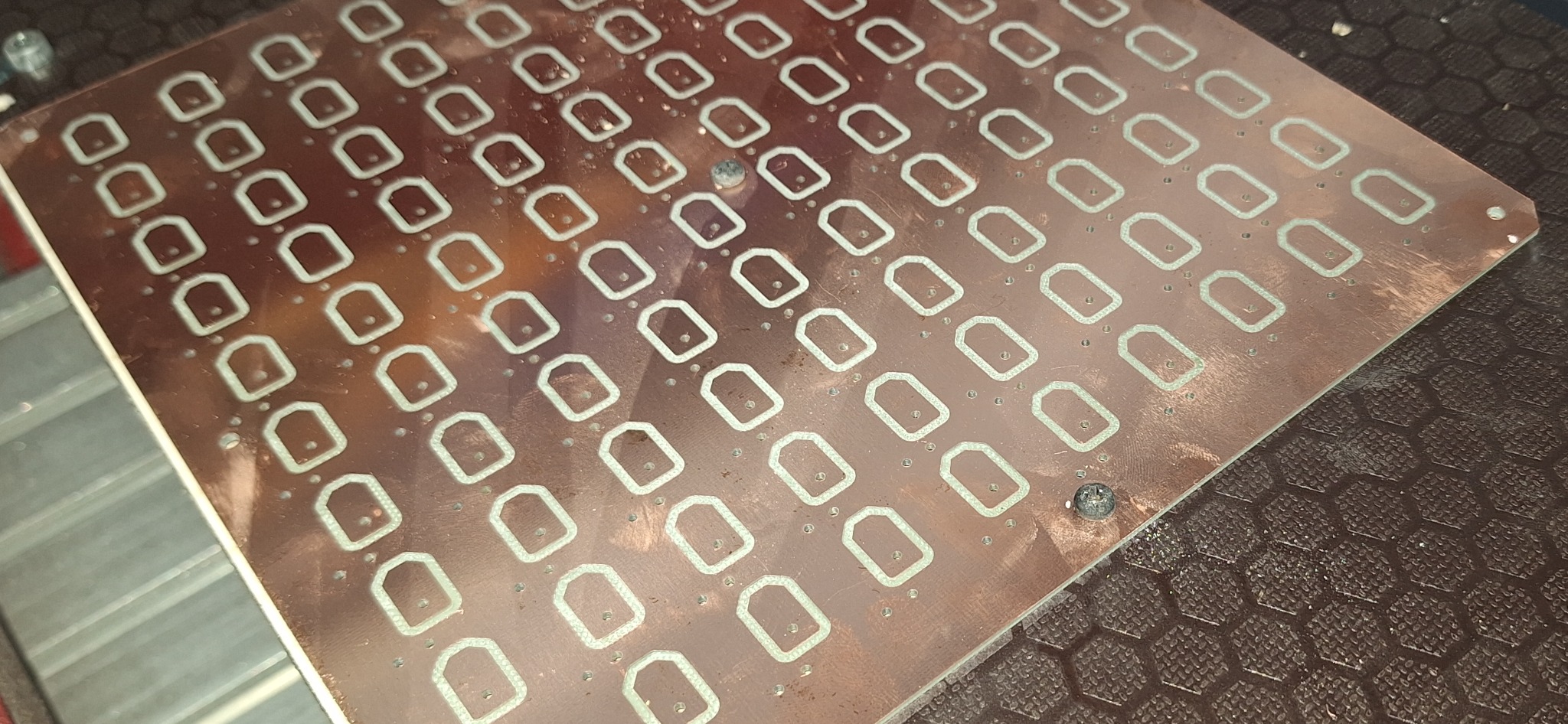 Miedziana płyta z wyfrezowanymi otworami w kształcie zaokrąglonych prostokątów i małymi otworami, przytwierdzona śrubami do podłoża.