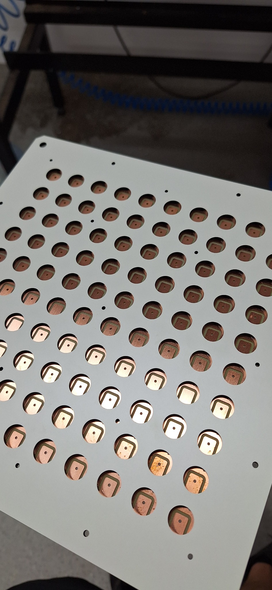 Szara płyta z regularnie rozmieszczonymi okrągłymi otworami, ukazującymi miedziane elementy z ciemnymi obwódkami i centralnymi punktami, widoczne ślady obróbki.