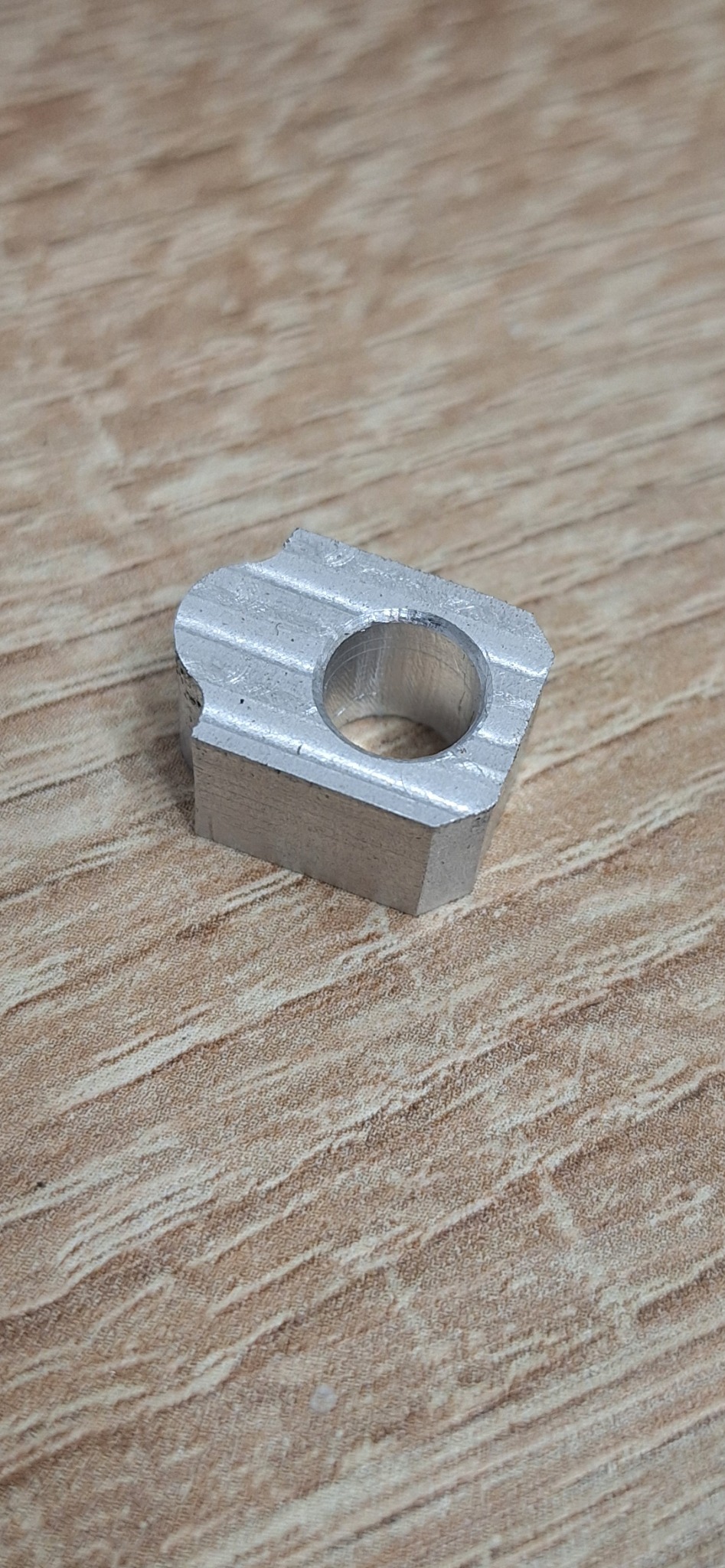 Szlifowany, aluminiowy element z otworem, leżący na drewnianej powierzchni, z widocznymi śladami obróbki.