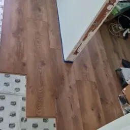 Panele podłogowe o wzorze drewna w trakcie układania, widoczne podkłady z nadrukowanymi liniami cięcia, niebieska taśma malarska przy ścianie, fragment niezabezpieczonej futryny drzwi.