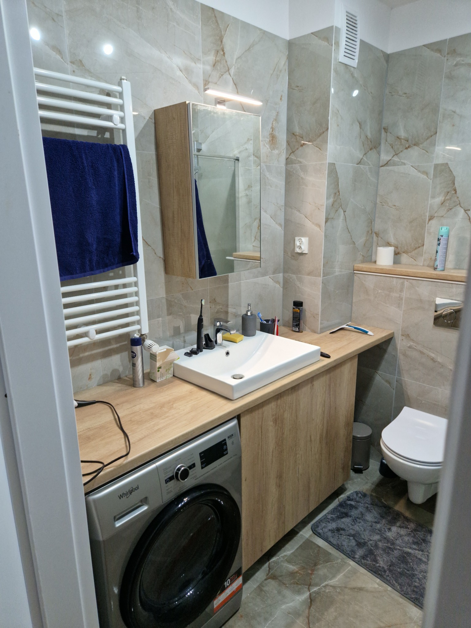 Wykończona łazienka z płytkami imitującymi marmur, pralką Whirlpool pod blatem umywalkowym, grzejnikiem drabinkowym z powieszonym granatowym ręcznikiem i wiszącą szafką z lustrem.