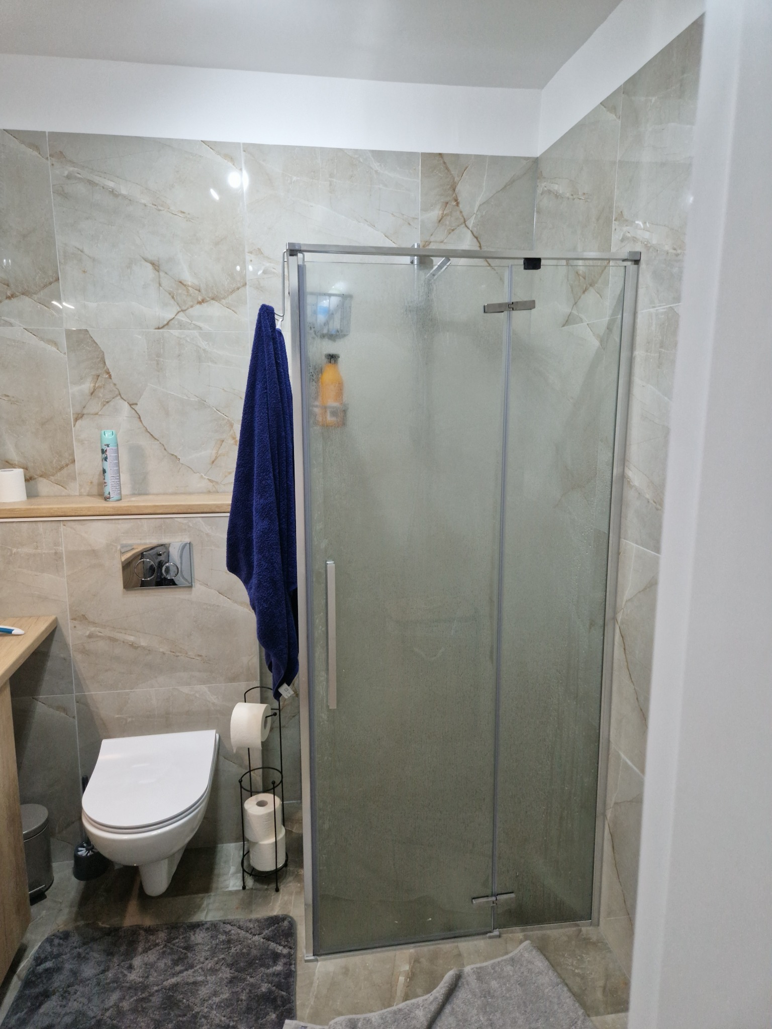Wykończona łazienka z kabiną prysznicową, beżowe marmurowe płytki na ścianach, wisząca toaleta i niebieski ręcznik.
